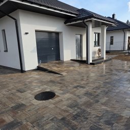 LexBud Oleksandr Pylypchuk - Ułożona kostka brukowa w odcieniach brązu i szarości na podjeździe przed nowoczesnym domem z garażem i czarnym dachem. Widoczny właz kanalizacyjny.