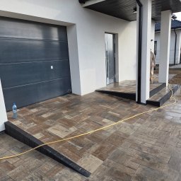 LexBud Oleksandr Pylypchuk - Ułożona kostka brukowa w odcieniach brązu i szarości przed nowoczesnym domem z garażem i kolumnami. Widoczny fragment węża ogrodowego i butelka wody.