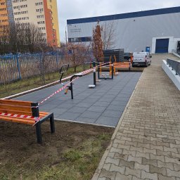 LexBud Oleksandr Pylypchuk - Nowa strefa aktywności fizycznej na świeżym powietrzu w Warszawie, wyłożona szarą kostką gumową, z ławkami i urządzeniami do ćwiczeń, otoczona taśmą ostrzegawczą.