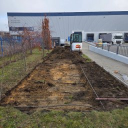LexBud Oleksandr Pylypchuk - Przygotowanie terenu pod kostkę brukową: wykopana ziemia, wytyczone granice, mini koparka Bobcat w tle, budynek magazynowy i ciężarówki. Widok z poziomu gruntu.