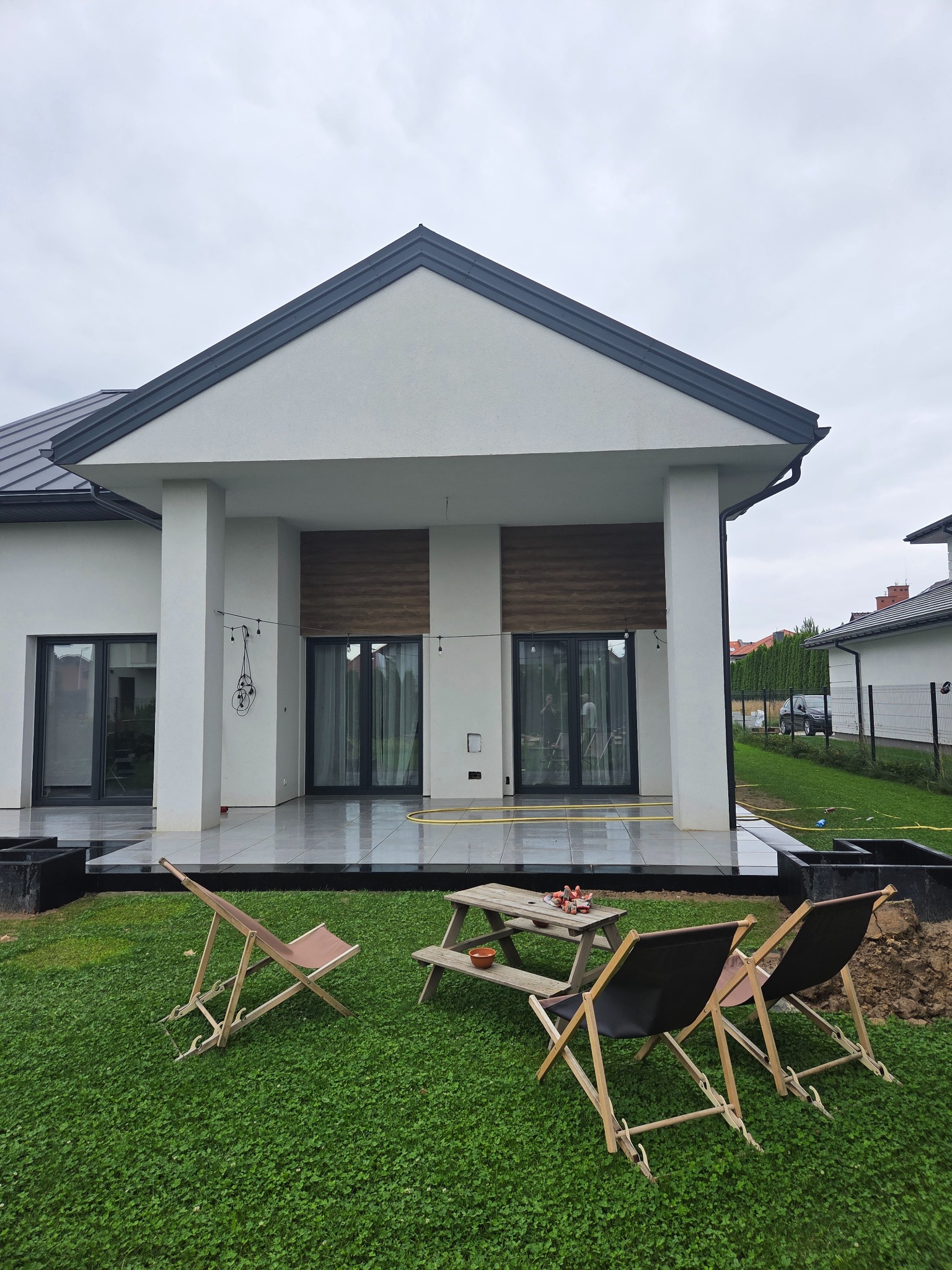 Nowoczesny taras z szarych płyt, drewniane detale elewacji, minimalistyczne meble ogrodowe na trawie. Widok na dom z płaskim dachem w pochmurny dzień.