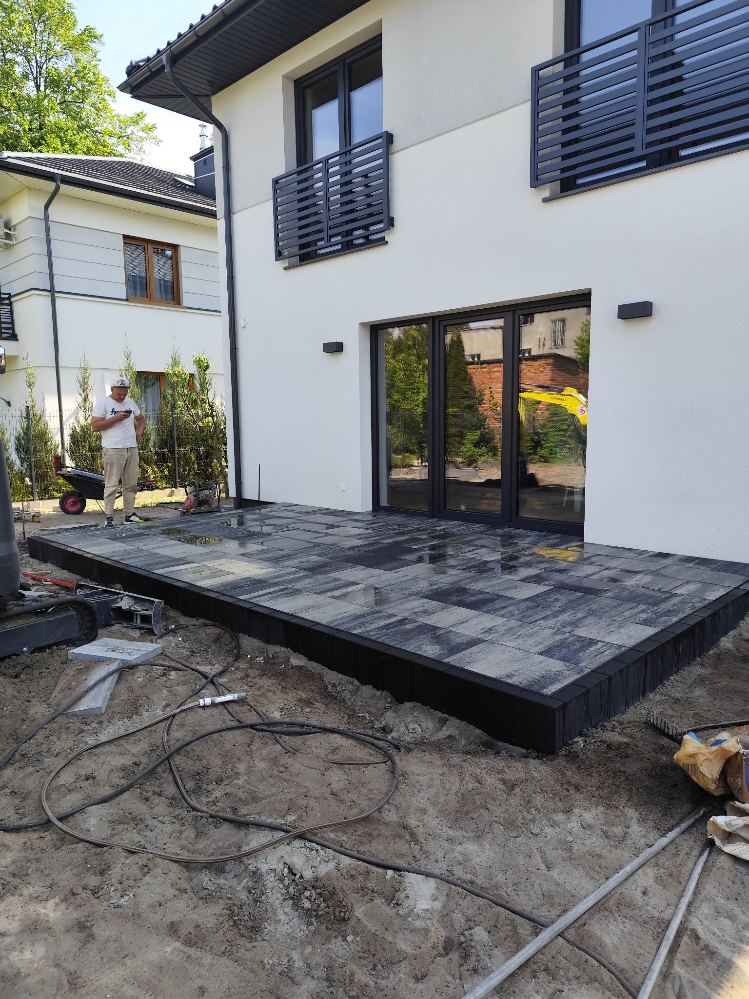 Nowoczesny taras z kostki brukowej w odcieniach szarości, z czarnym obrzeżem, przylegający do jasnego budynku z dużymi oknami. W tle pracownik i sprzęt budowlany.