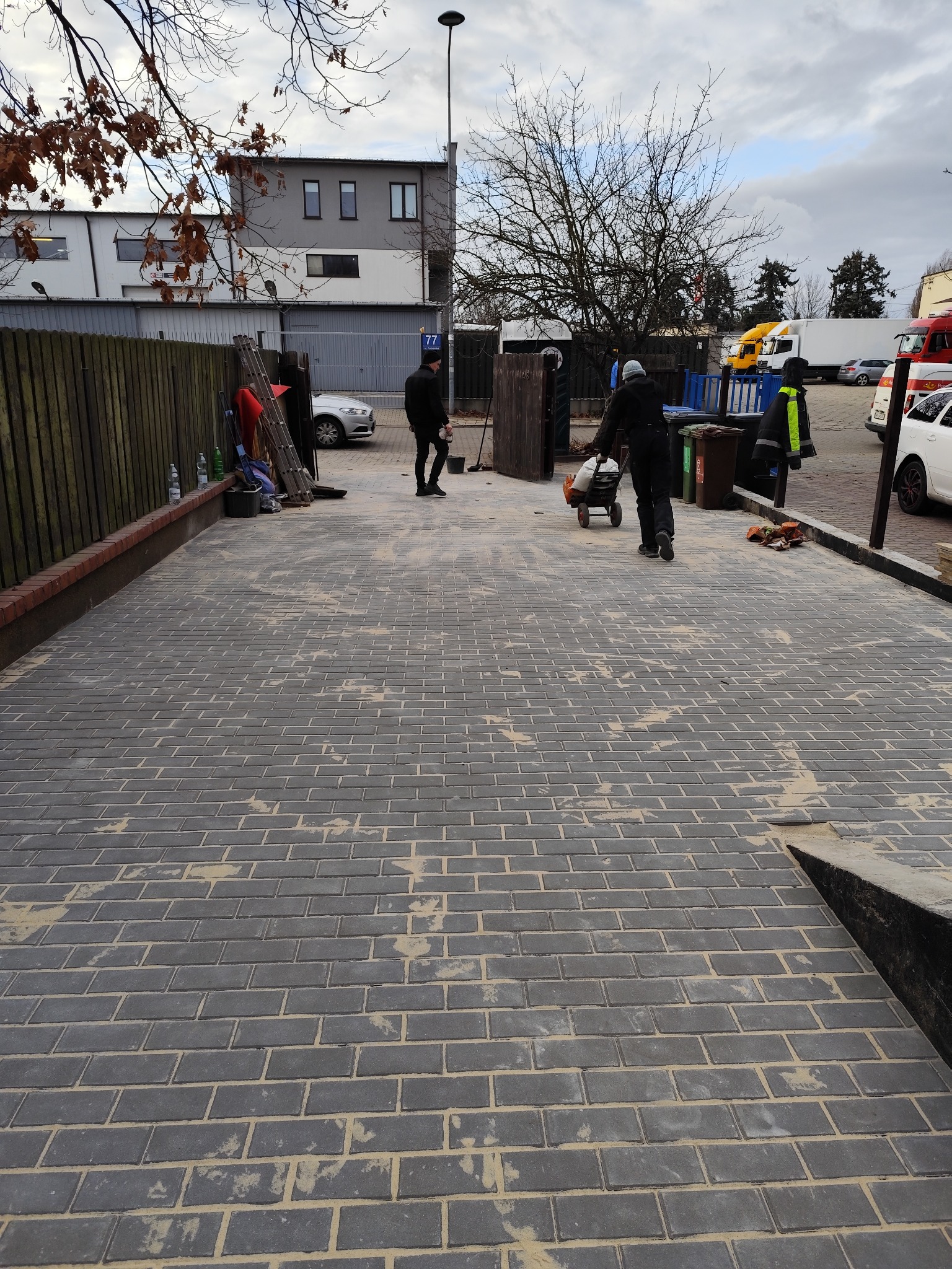 Ułożona szara kostka brukowa na podjeździe, z elementami piasku w spoinach. Pracownicy i sprzęt w tle sugerują trwające prace wykończeniowe.