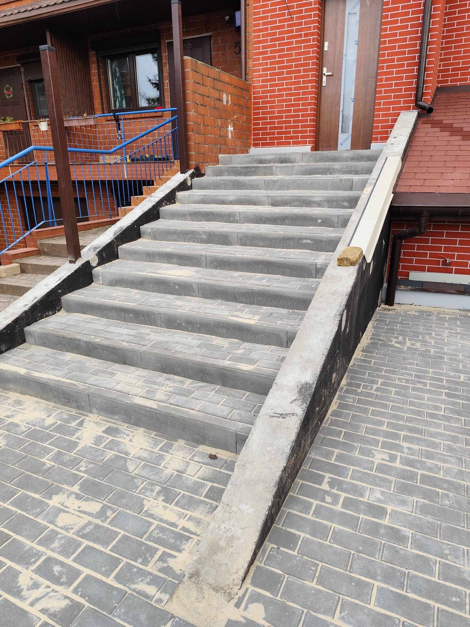 Schody i chodnik z szarej kostki brukowej przed domem z czerwonej cegły. Widoczny fragment dachu i rynny. Kostka posypana piaskiem.
