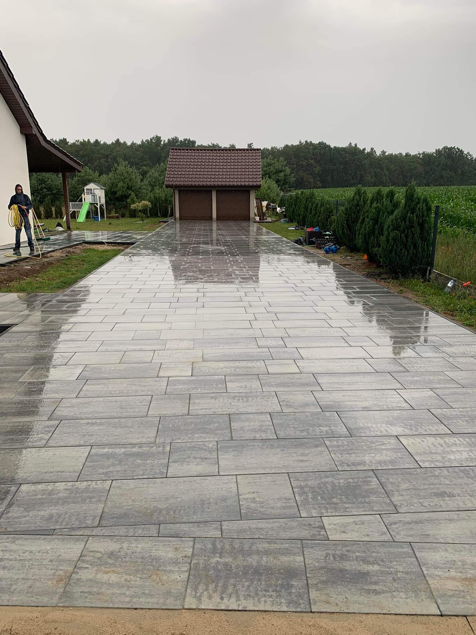 Ułożona, mokra kostka brukowa w odcieniach szarości prowadząca do garażu. W tle zieleń i dom z osobą z wężem ogrodowym. Widok na posesję w pochmurny dzień.