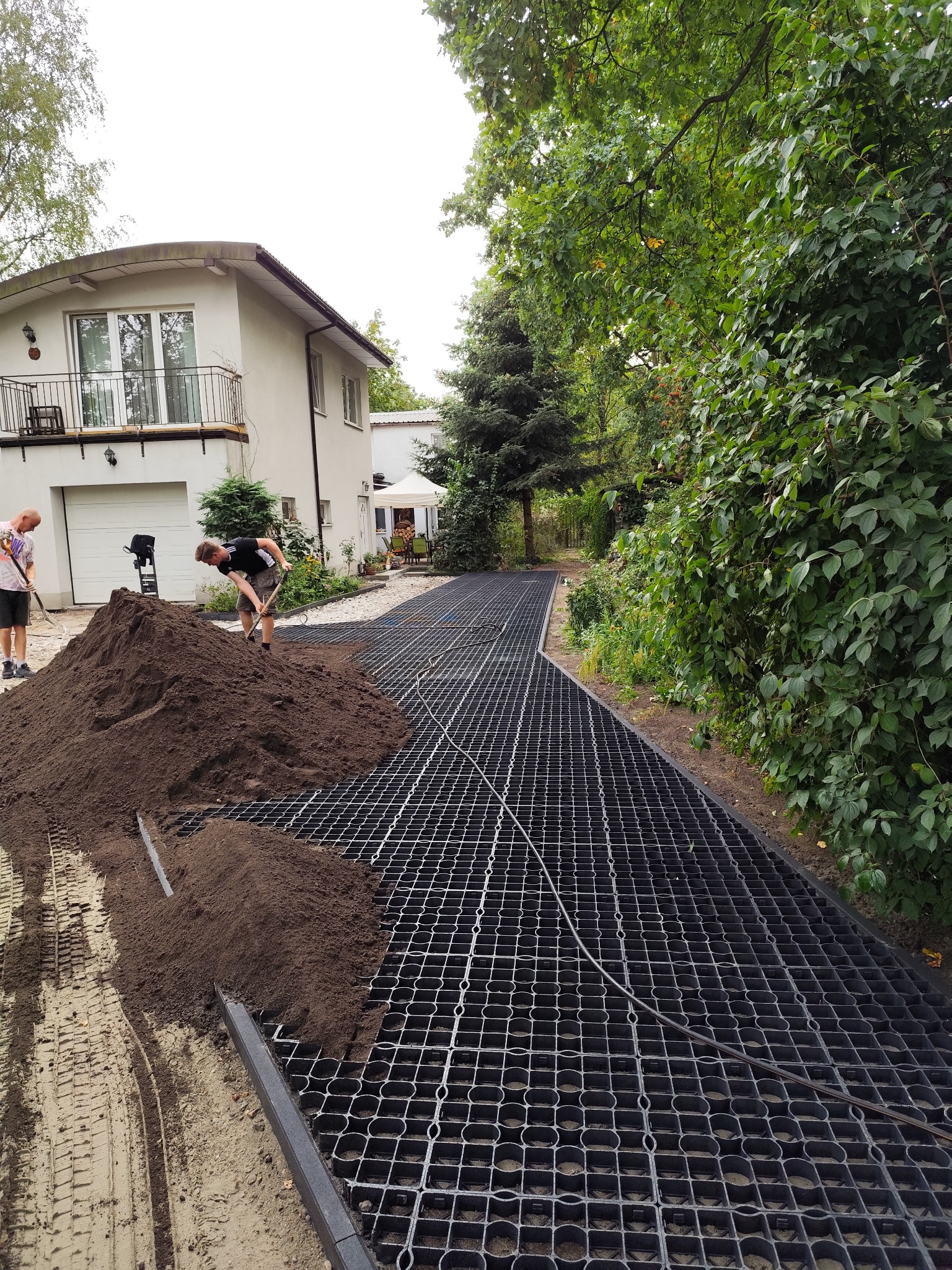 Przygotowanie podłoża: robotnicy rozgarniają ziemię na plastikowej siatce, tworząc podbudowę pod nawierzchnię na tle domu z garażem.