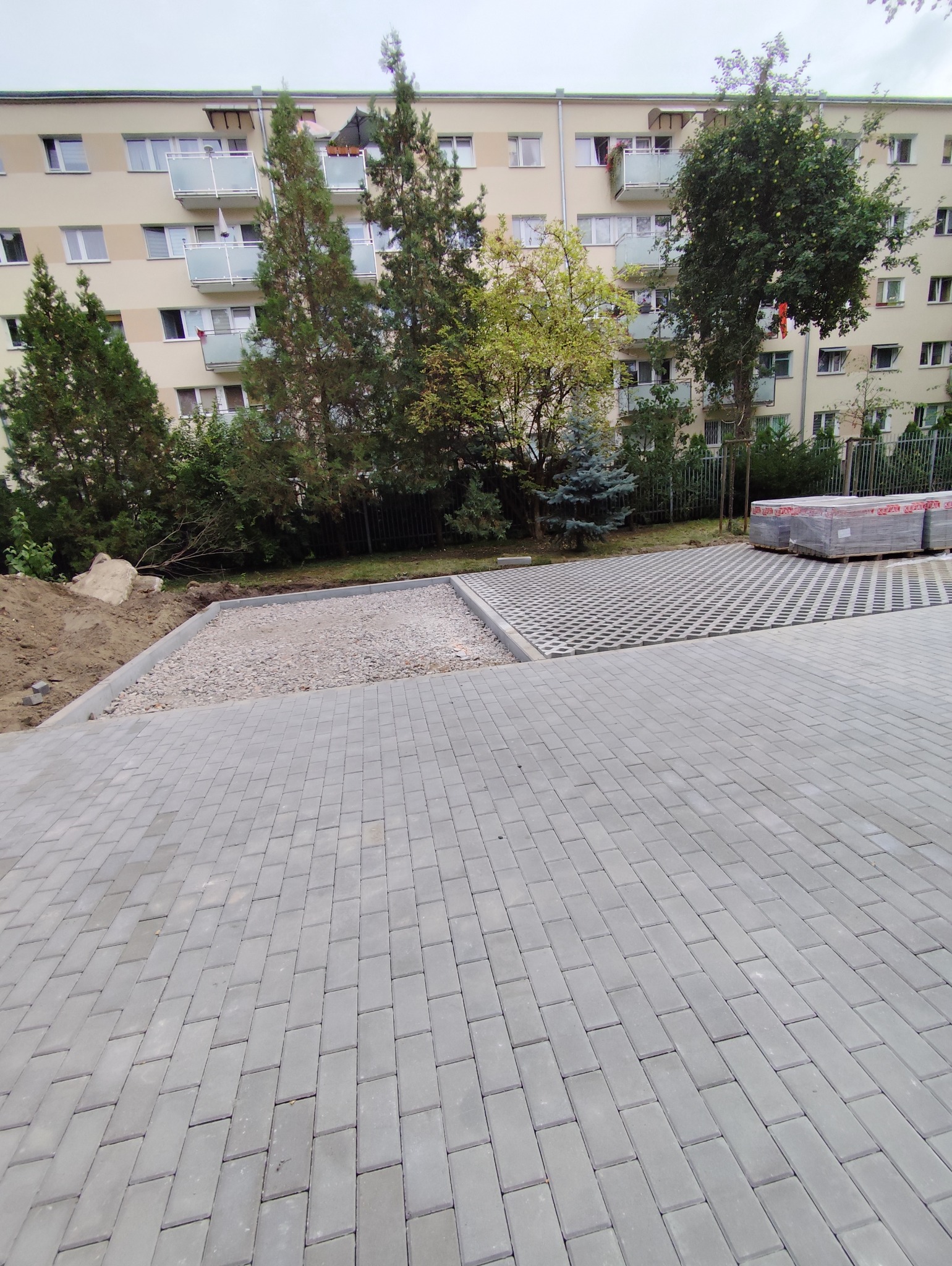 Częściowo ułożona szara kostka brukowa na podjeździe przed blokiem mieszkalnym. Widoczny fragment podbudowy z kruszywa i paleta kostki.