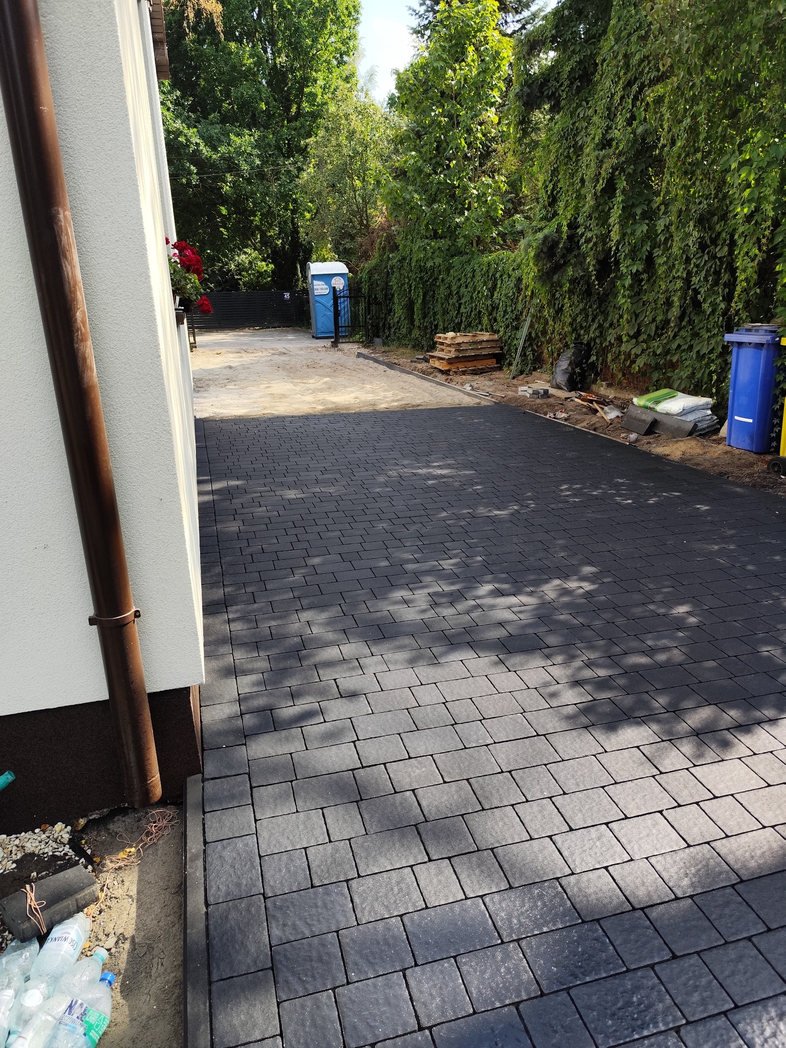 Ciemnoszara kostka brukowa ułożona na podjeździe, z widocznymi elementami ogrodu i fragmentem budynku. Na końcu podjazdu widoczna toaleta przenośna.