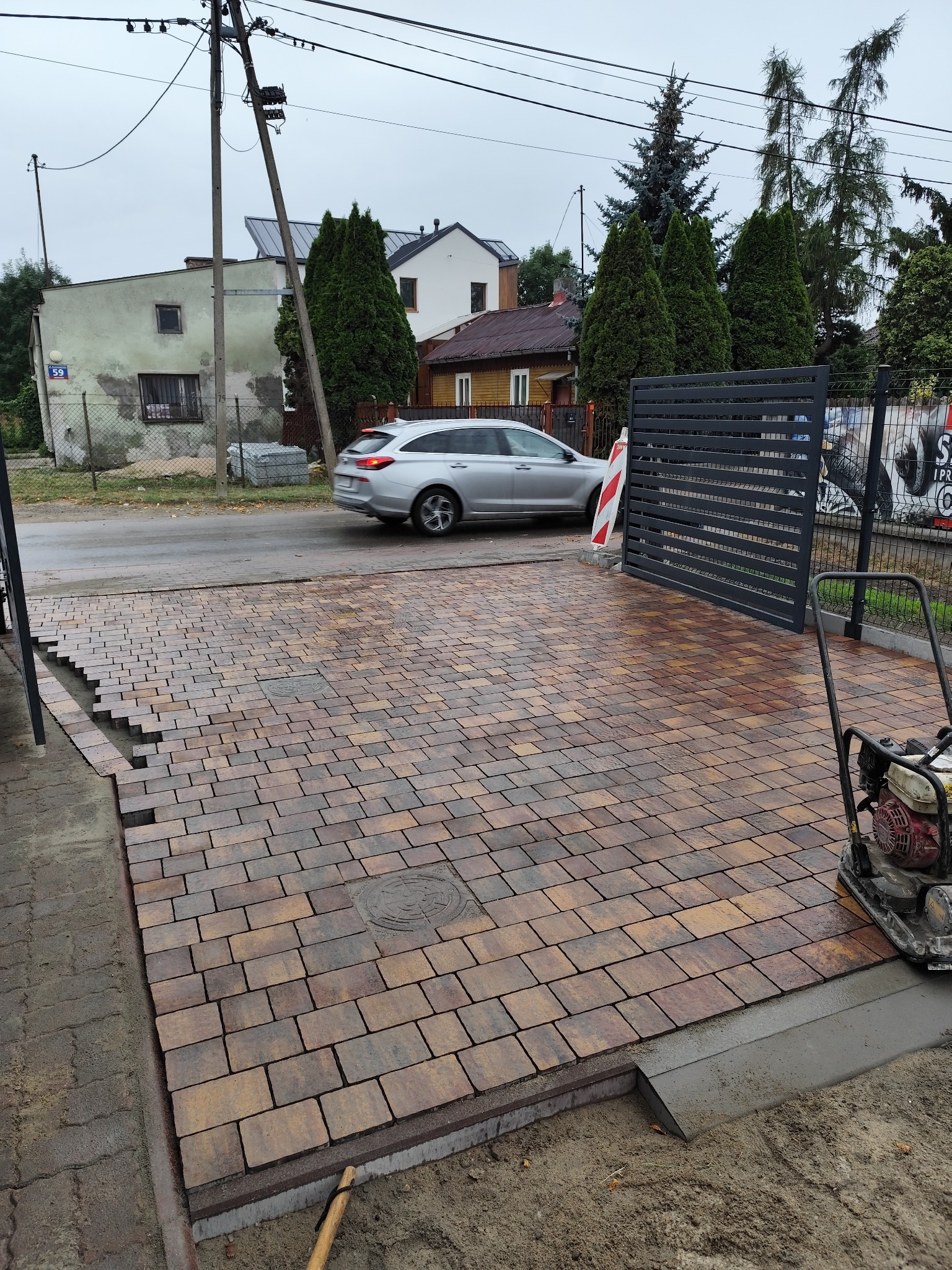 Częściowo ułożona, brązowo-pomarańczowa kostka brukowa na podjeździe, z zagęszczarką w tle. Widoczny fragment ulicy z budynkami i samochodem osobowym.