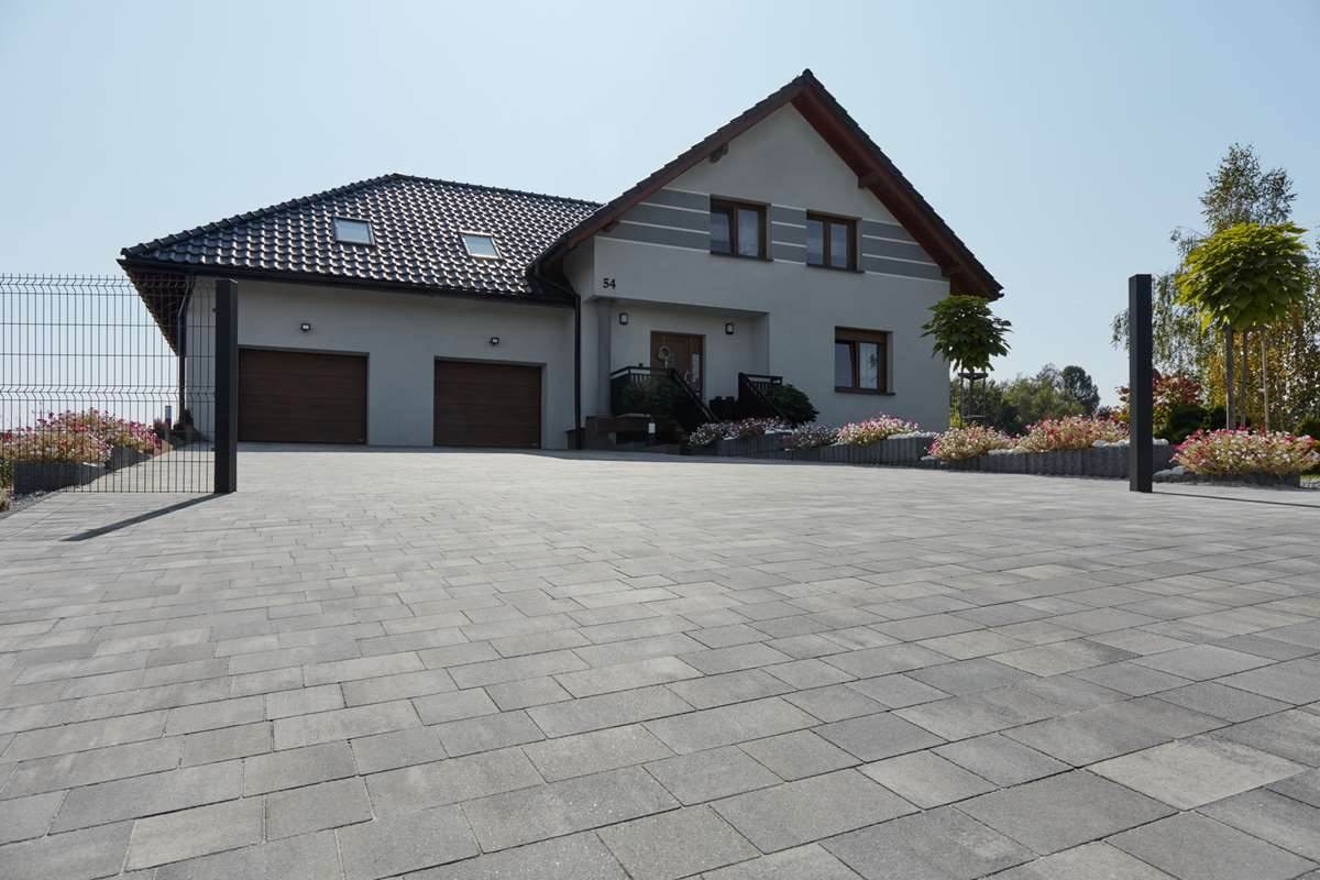Szara kostka brukowa na podjeździe do domu z dwoma garażami, rabaty kwiatowe oddzielają podjazd od budynku. Widoczne ogrodzenie z siatki.