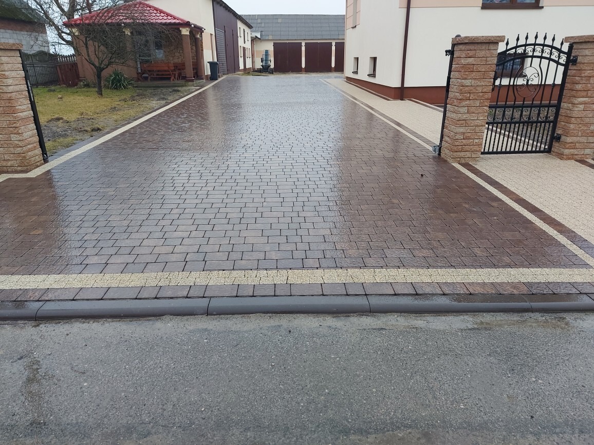 Brązowa kostka brukowa ułożona na podjeździe, wykończona jasnymi pasami i szarymi obrzeżami. W tle budynki i brama wjazdowa. Po deszczu, mokra nawierzchnia.