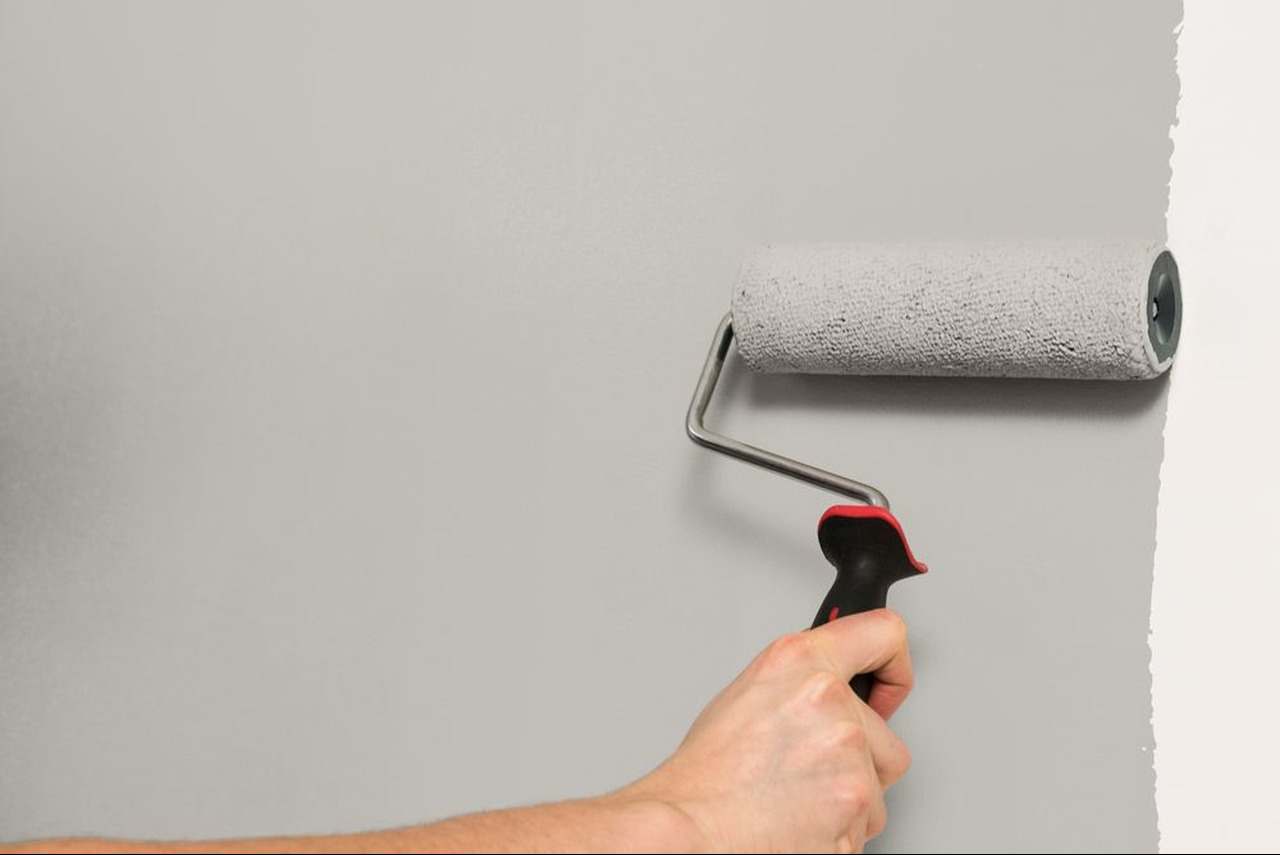 Ręka z wałkiem malarskim nanosi szarą farbę na ścianę, widoczna granica świeżo pomalowanej powierzchni.