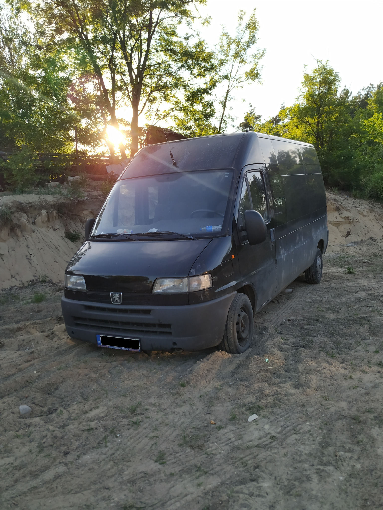 Czarny, lekko zabrudzony furgon Peugeot zaparkowany na piaszczystym terenie z widocznymi śladami opon, w tle drzewa i zachodzące słońce.