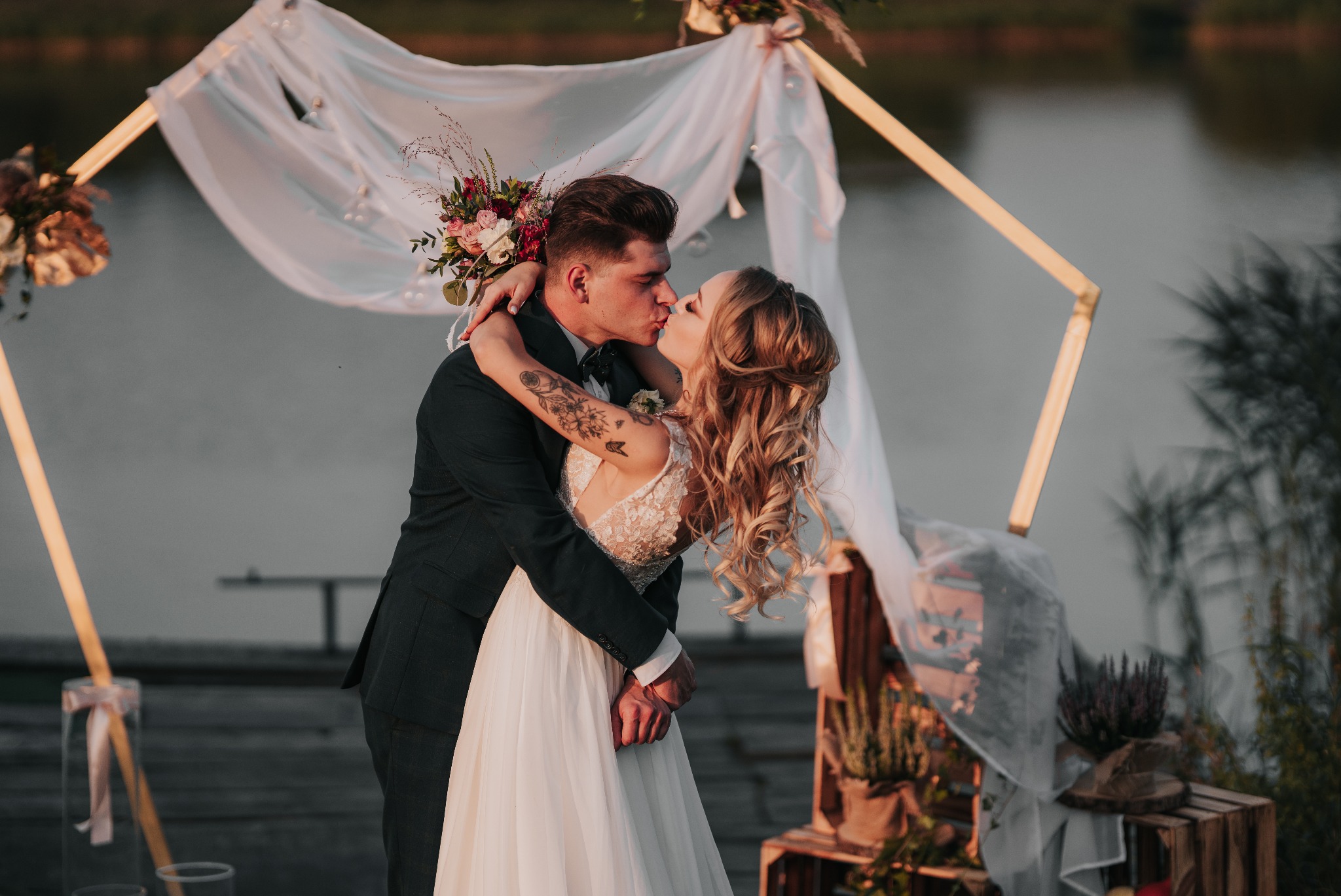 Pocałunek młodej pary pod dekoracyjnym, geometrycznym łukiem z białej tkaniny i kwiatów, na tle jeziora i zieleni.