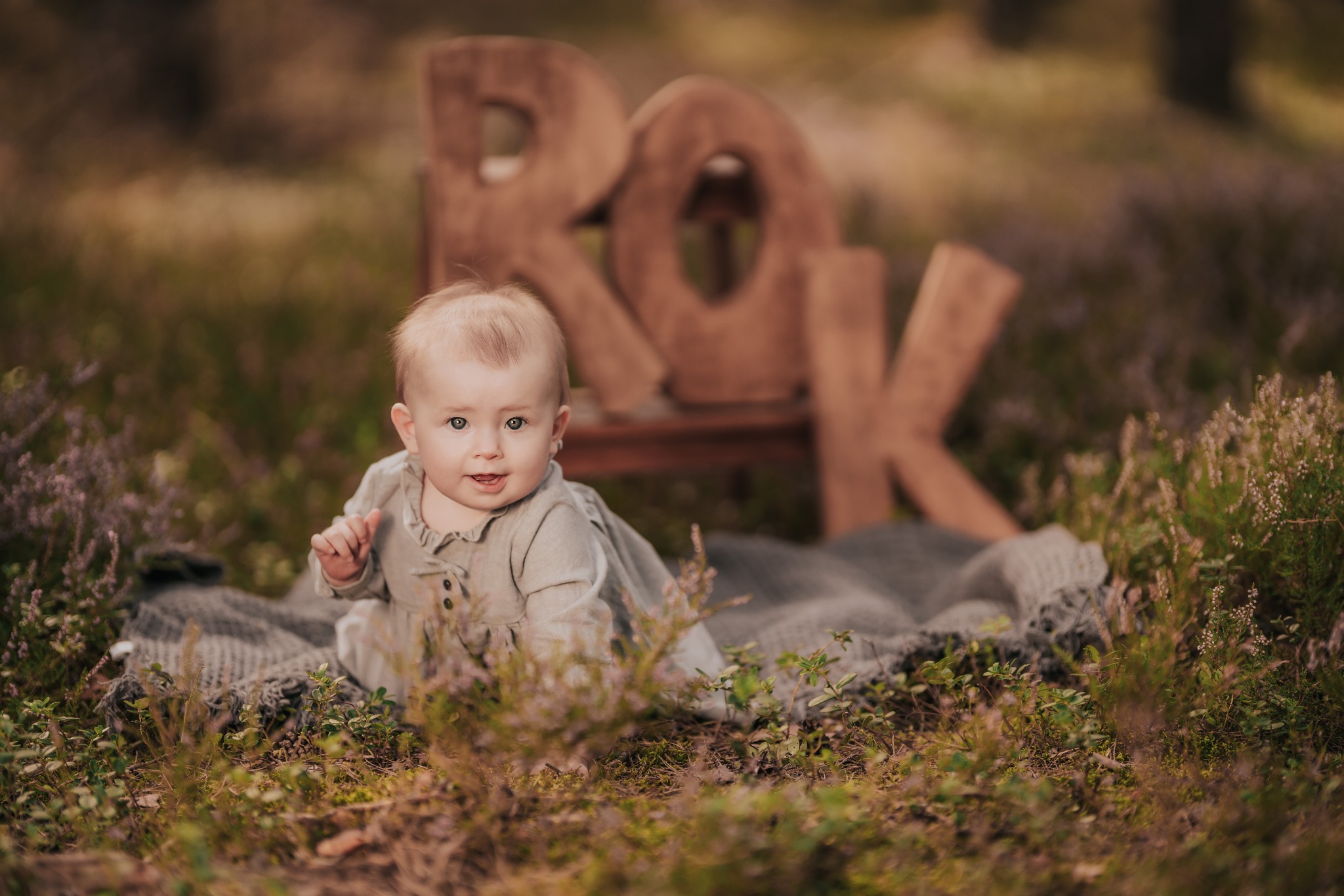 Urocze niemowlę z błyszczącymi oczami w Kluczborku, siedzące na kocu w lesie, z drewnianym napisem 'ROK' w tle.