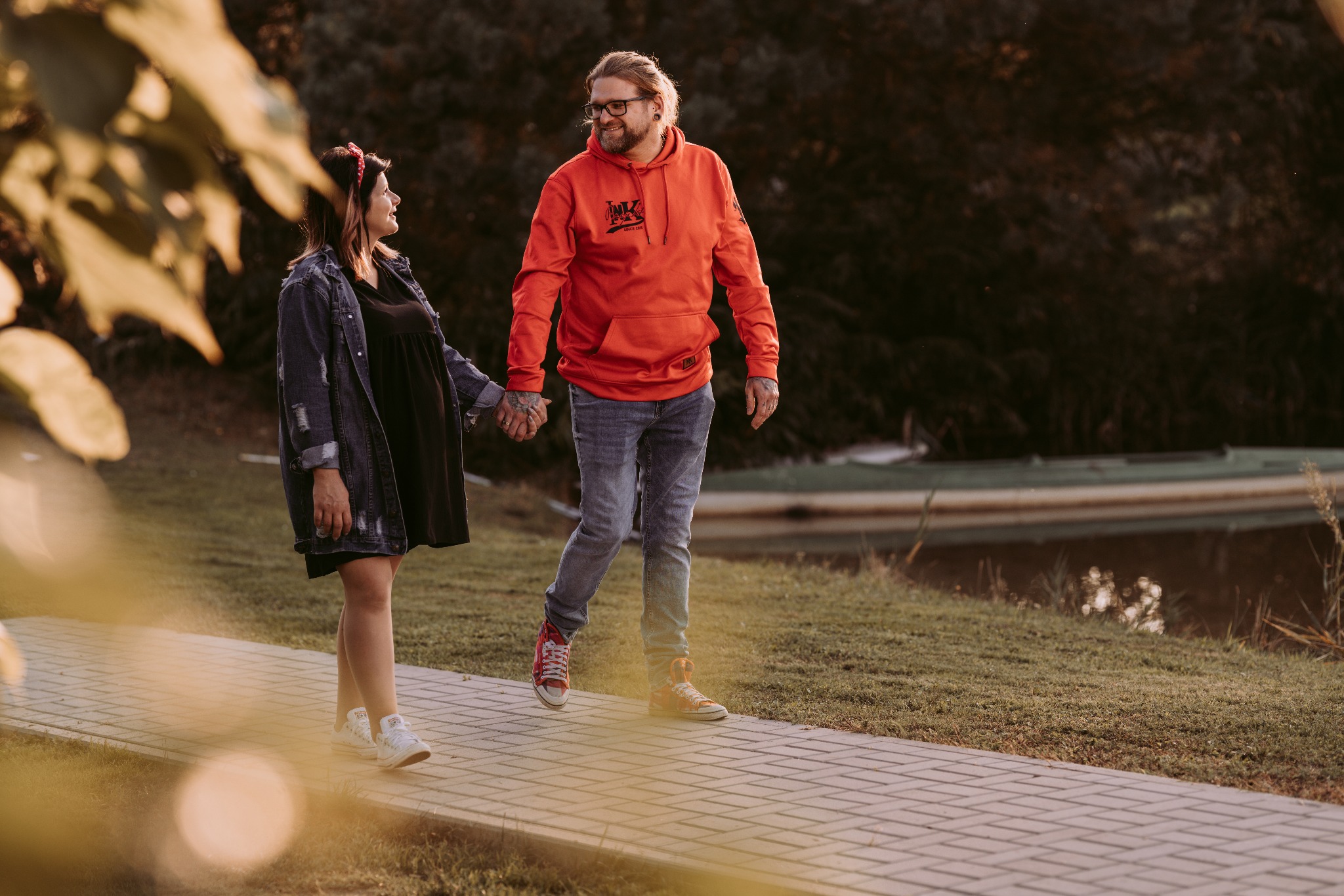 Uśmiechnięta para trzymająca się za ręce spaceruje po ścieżce w parku, kobieta w czarnej sukience i dżinsowej kurtce, mężczyzna w pomarańczowej bluzie z kapturem i dżinsach, w tle widać zarośnięte...