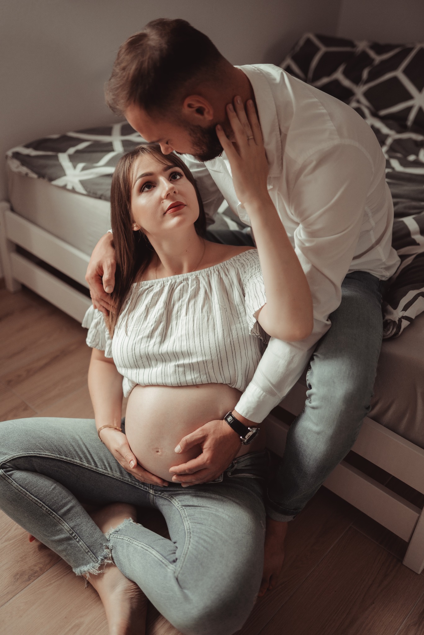 Ciężarna kobieta w bluzce w paski i jeansach siedzi na podłodze, obejmowana przez mężczyznę w białej koszuli i jeansach, który opiera głowę o jej czoło. Ona patrzy na niego z uśmiechem.