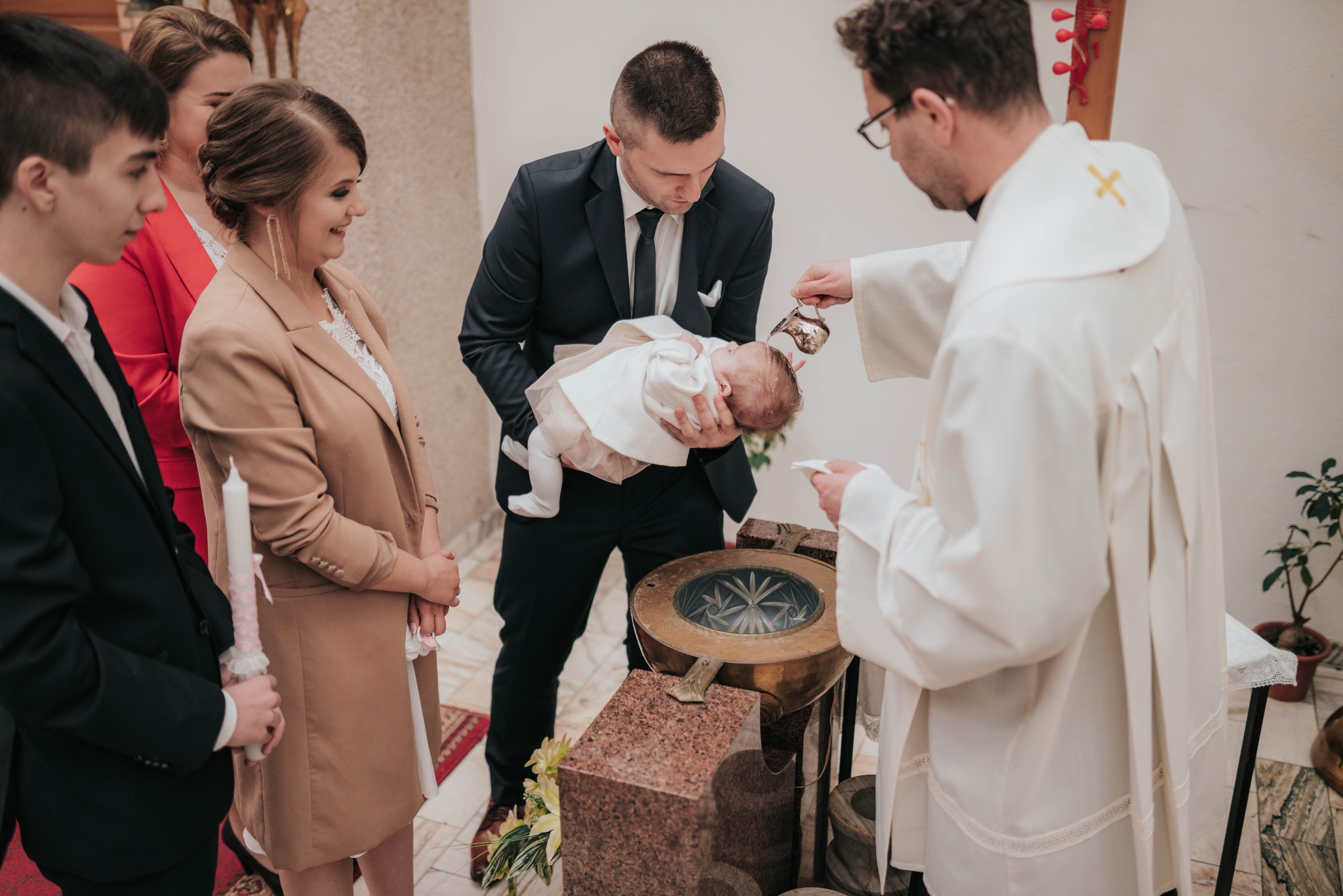 Uroczystość chrztu: ksiądz polewa wodą główkę niemowlęcia trzymanego przez ojca, w tle rodzina z świecami, w kościele.