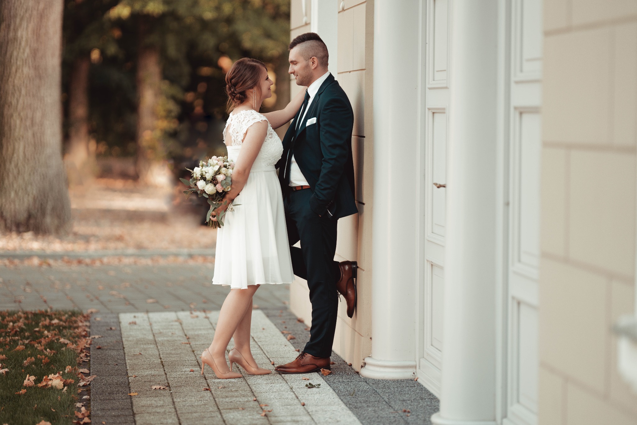 Para młoda stoi oparta o kolumnę budynku, panna młoda trzyma bukiet, oboje patrzą na siebie.