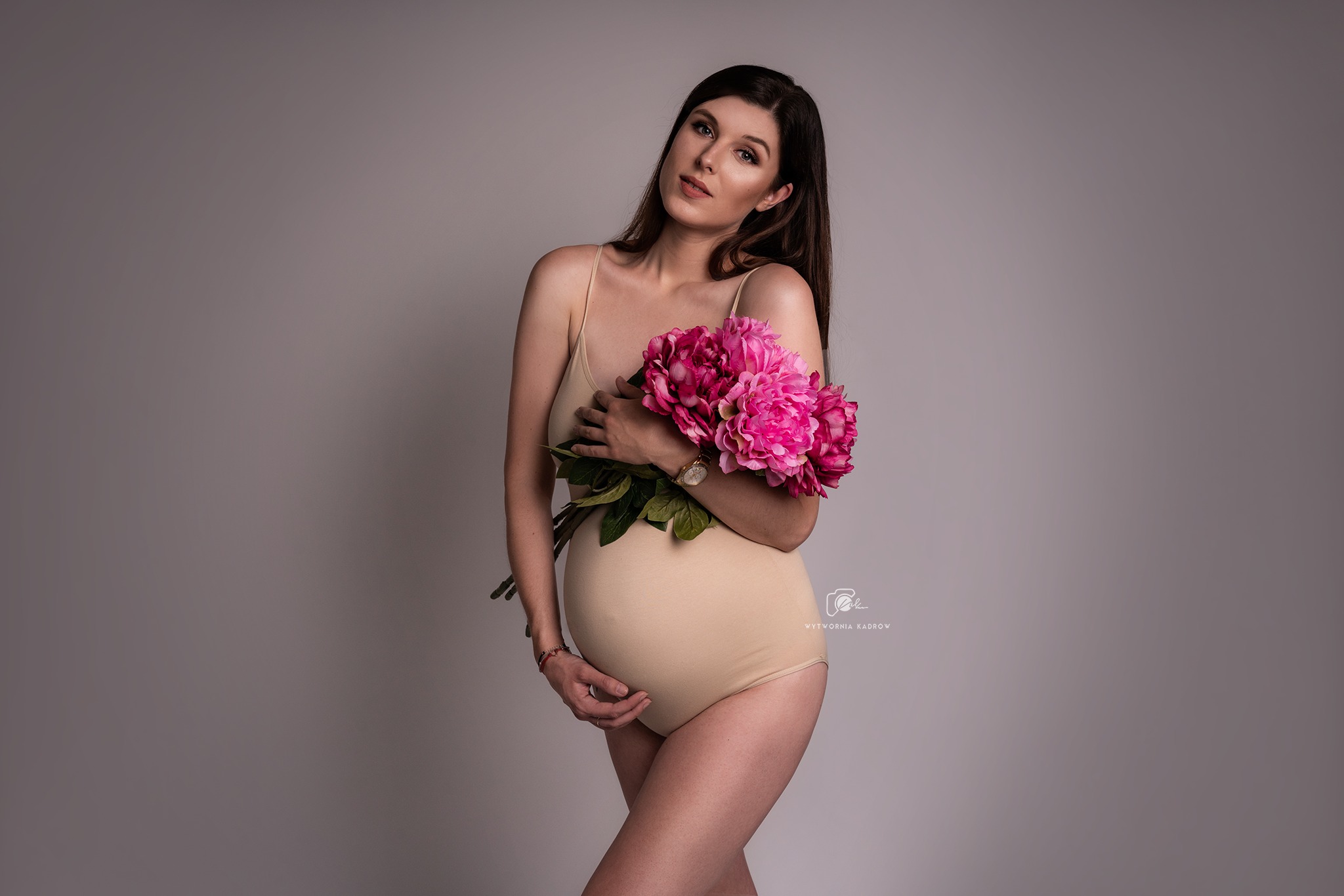 Kobieta w beżowym body, w ciąży, pozuje z bukietem różowych piwonii na szarym tle, delikatny makijaż, widoczny zegarek na ręce.