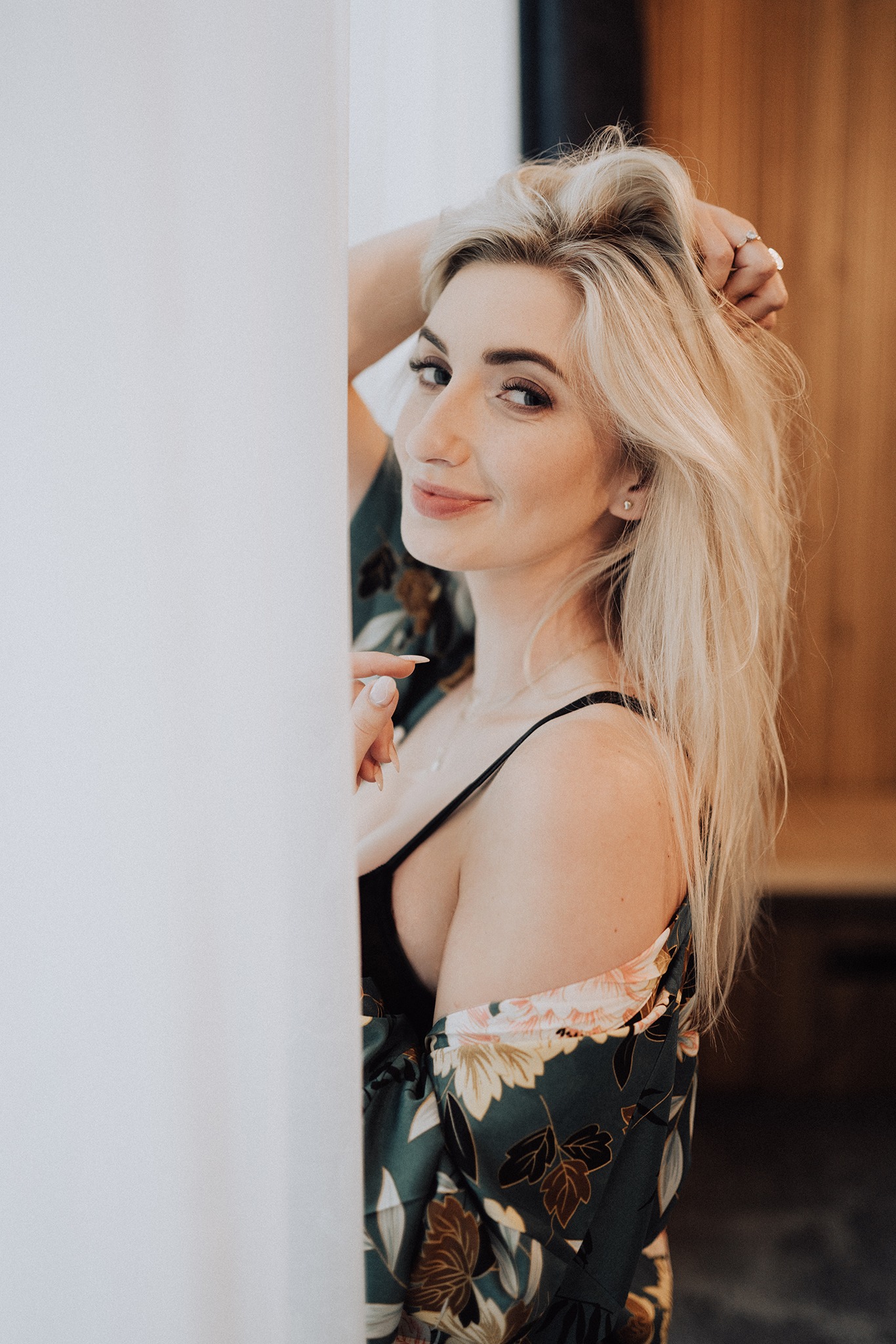 Uśmiechnięta blondynka w kwiecistym szlafroku, opierająca się o białą ścianę, z pierścionkiem na palcu i delikatnym makijażem.