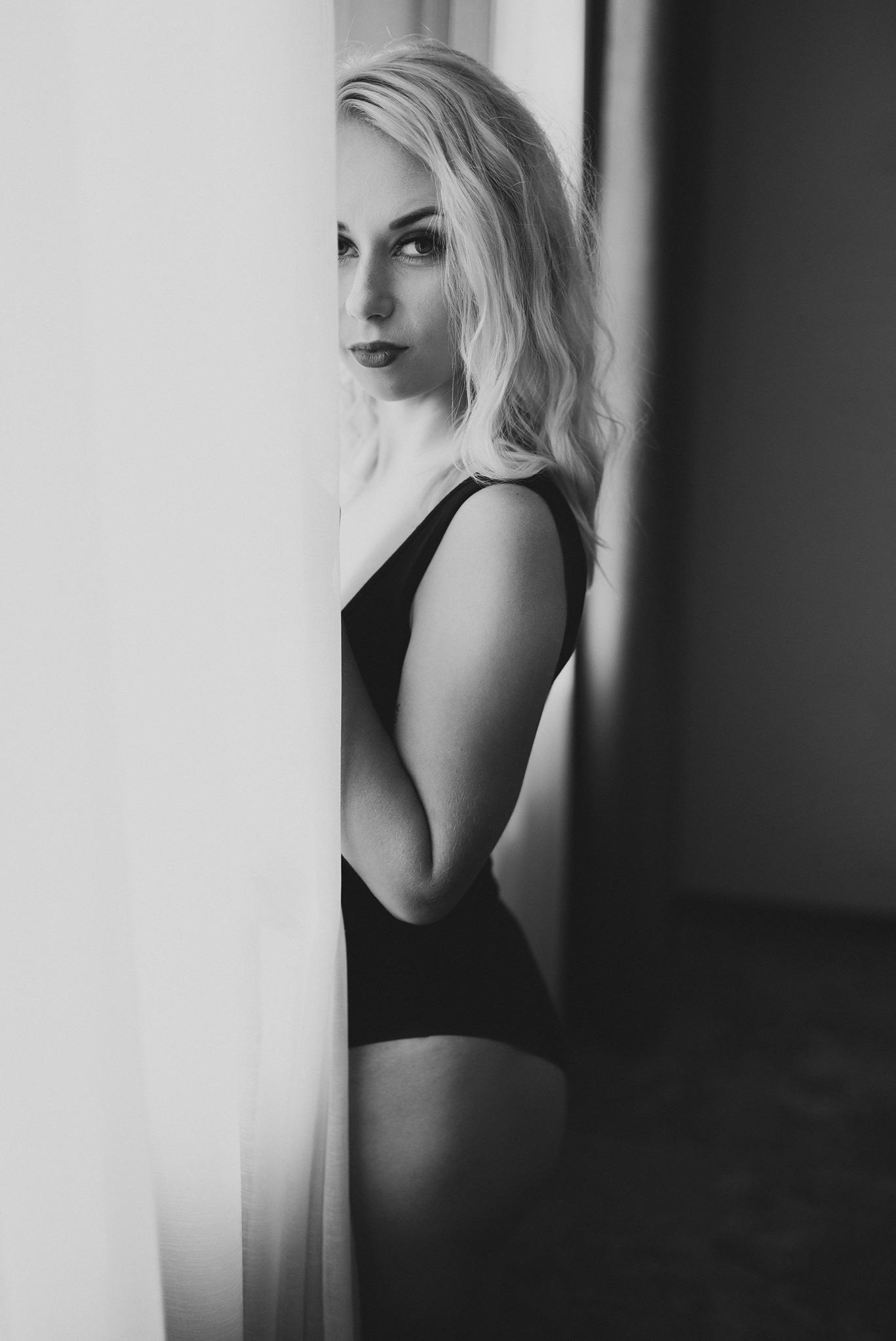Czarno-biały portret kobiety z blond włosami, w czarnym body, stojącej przy oknie, z intensywnym makijażem ust i spojrzeniem w obiektyw.