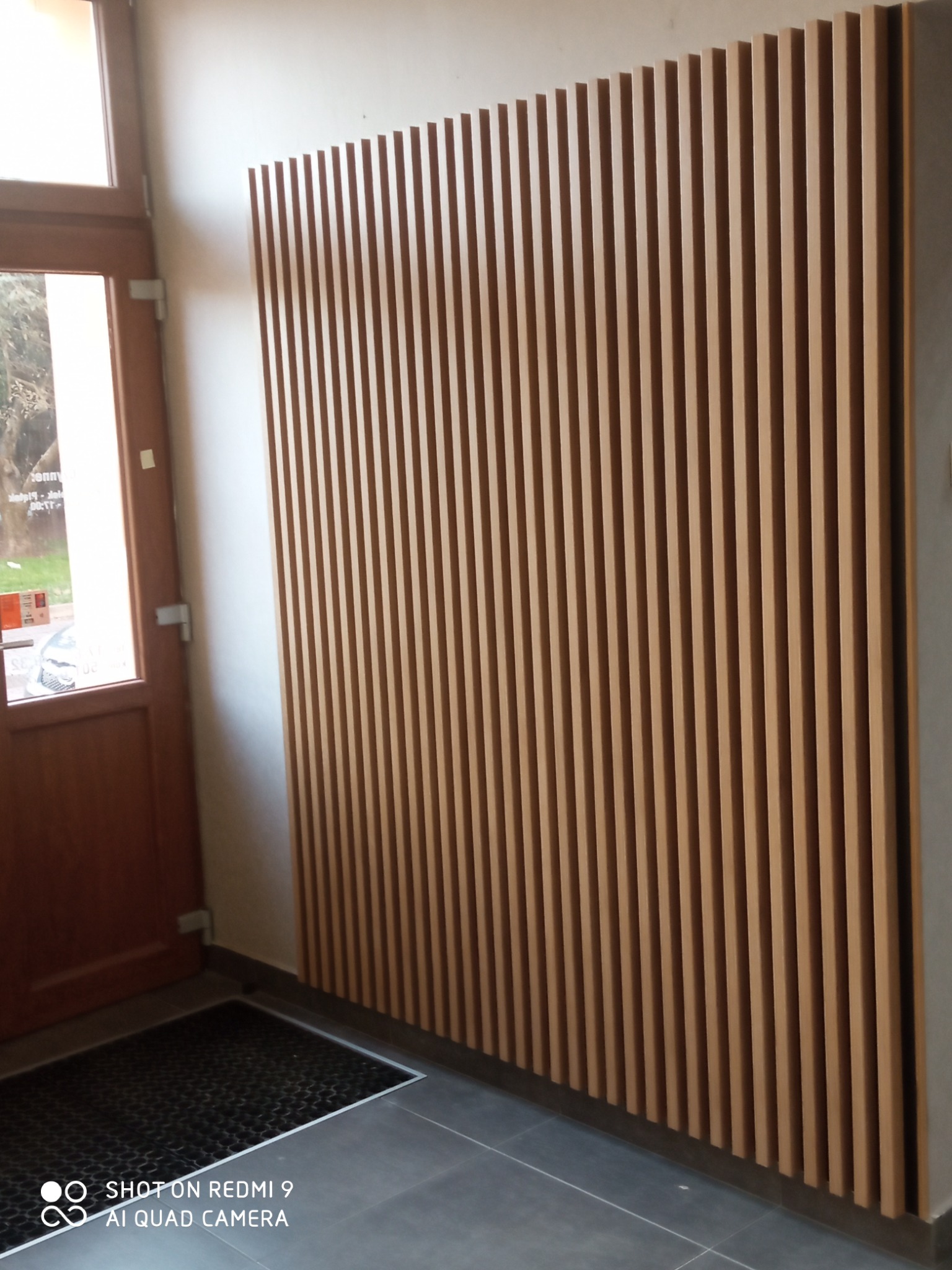 Panel dekoracyjny z pionowych listew drewnianych na ścianie w korytarzu, widoczny fragment drzwi wejściowych i wycieraczka.