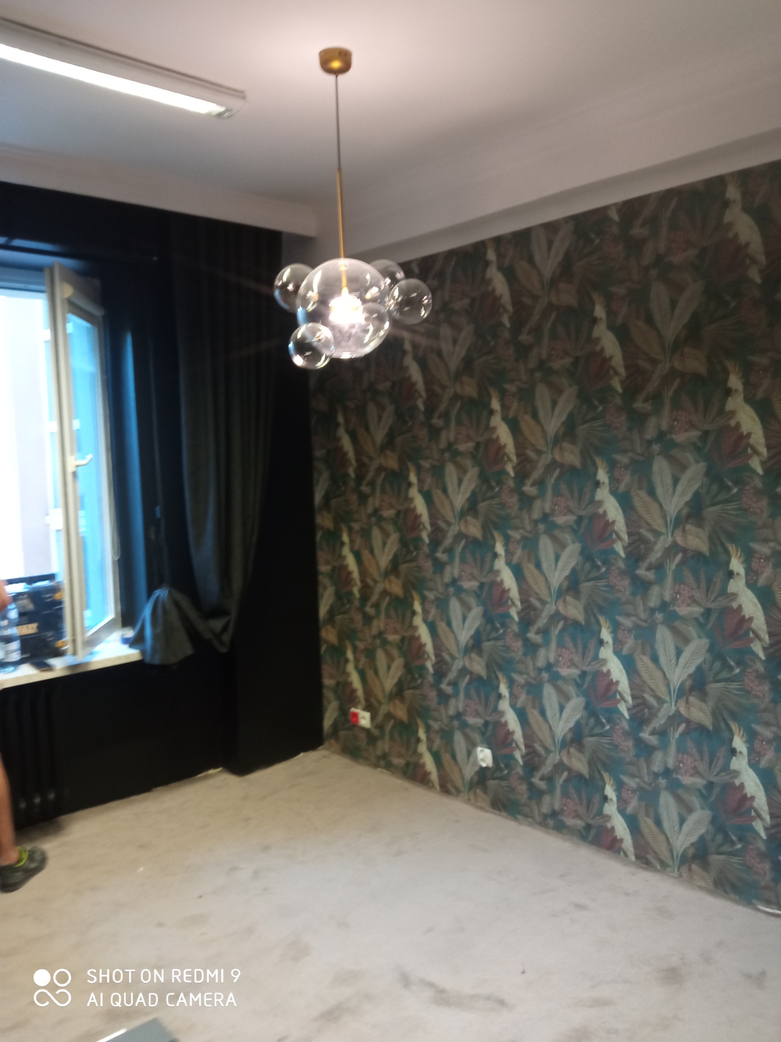 Pokój po remoncie z tapetą w roślinny wzór z motywem kakadu, widoczne okno z czarnymi zasłonami i nowoczesna lampa sufitowa.