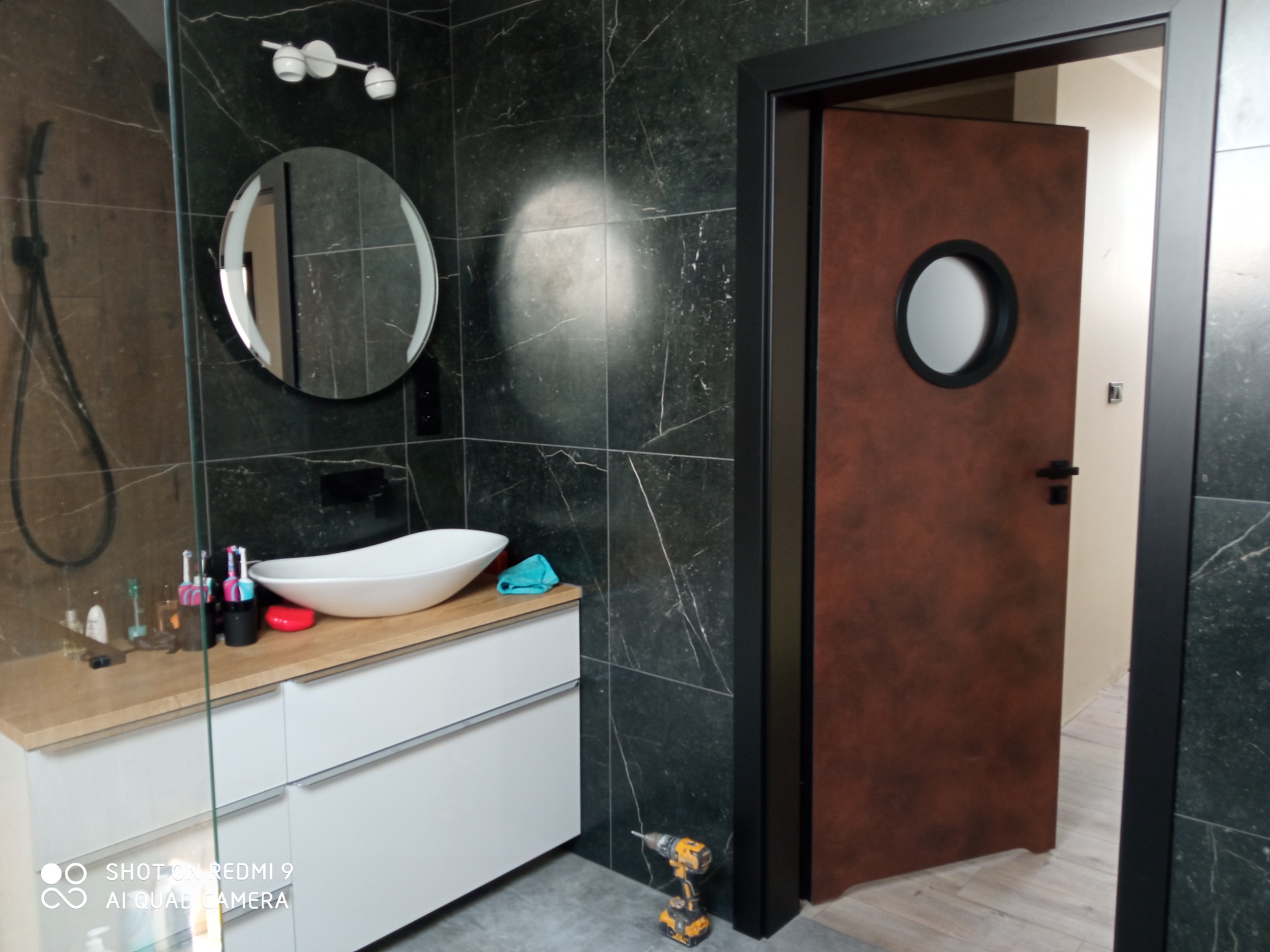 Nowoczesna łazienka z czarnymi marmurowymi płytkami, białą umywalką nablatową i brązowymi drzwiami z okrągłym okienkiem. W rogu widać wiertarkę akumulatorową.