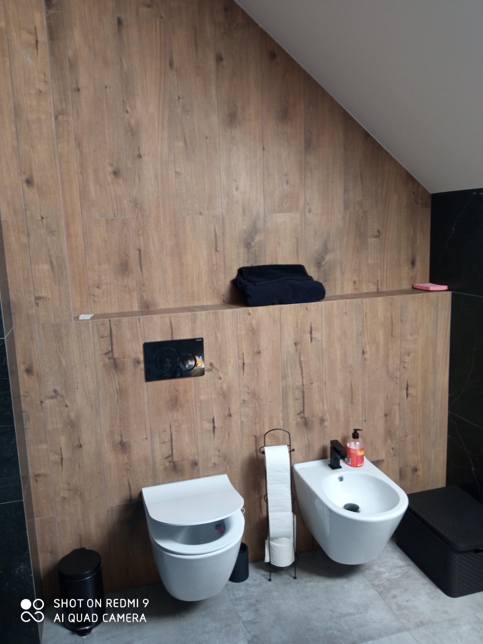 Nowoczesna łazienka w skandynawskim stylu z drewnopodobnymi płytkami na ścianach, wiszącą toaletą i bidetem z czarną armaturą, uzupełniona o czarne dodatki i półkę z ręcznikiem.