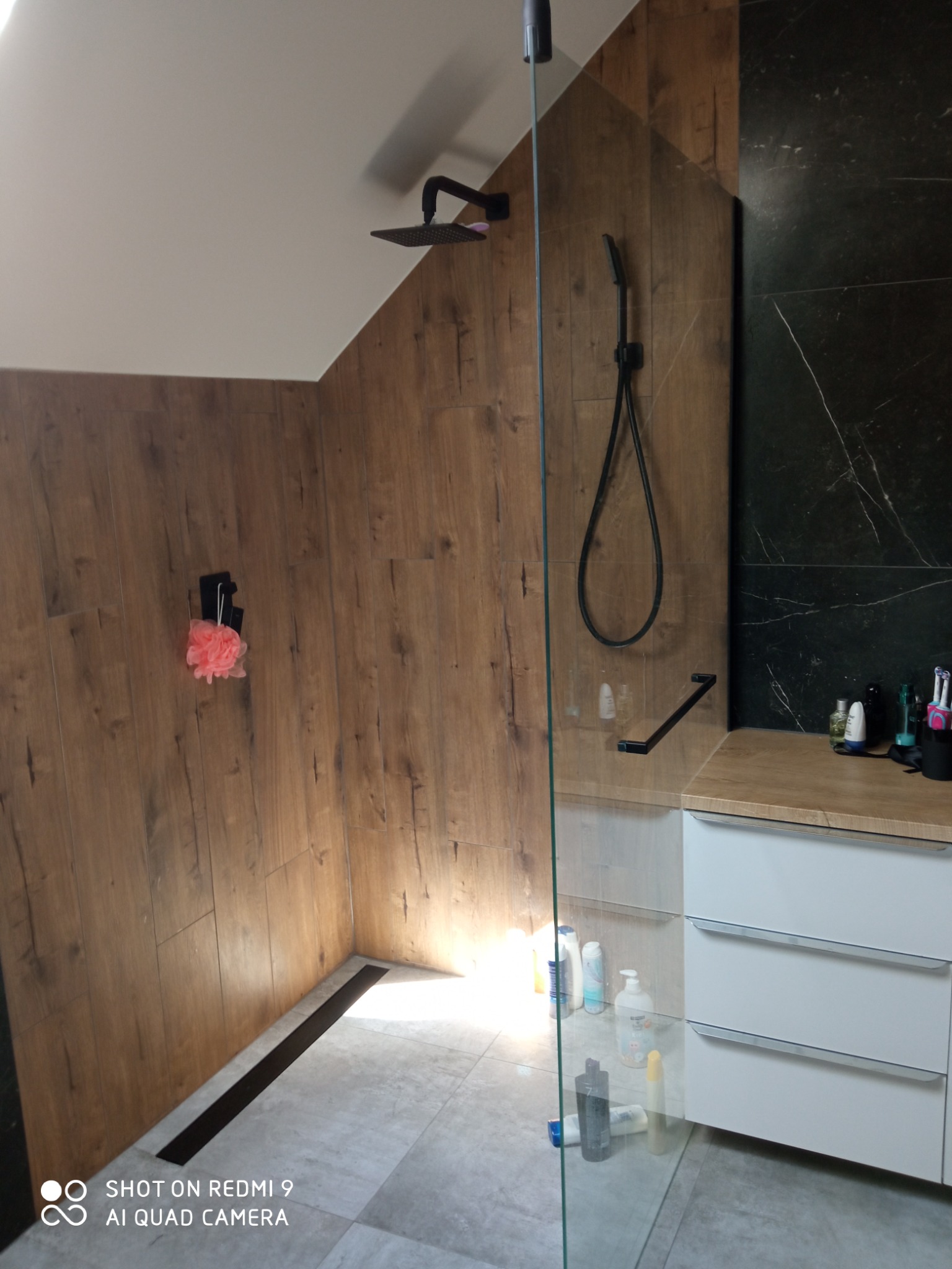 Nowoczesna kabina prysznicowa z drewnopodobnymi panelami na ścianach, czarną armaturą i odpływem liniowym, widoczna przez szklaną ściankę.