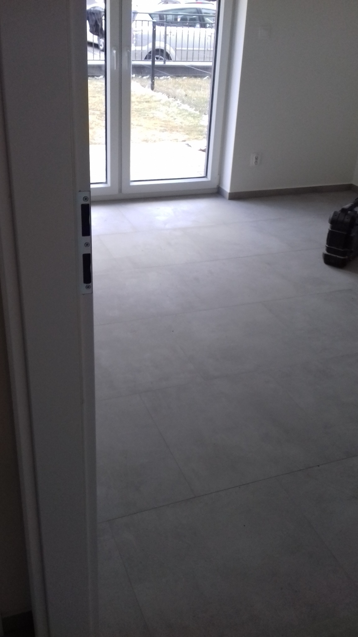 Otwarta biała rama drzwiowa z widokiem na jasne, nowo wyłożone płytkami podłogi i duże okno balkonowe z widokiem na posesję.