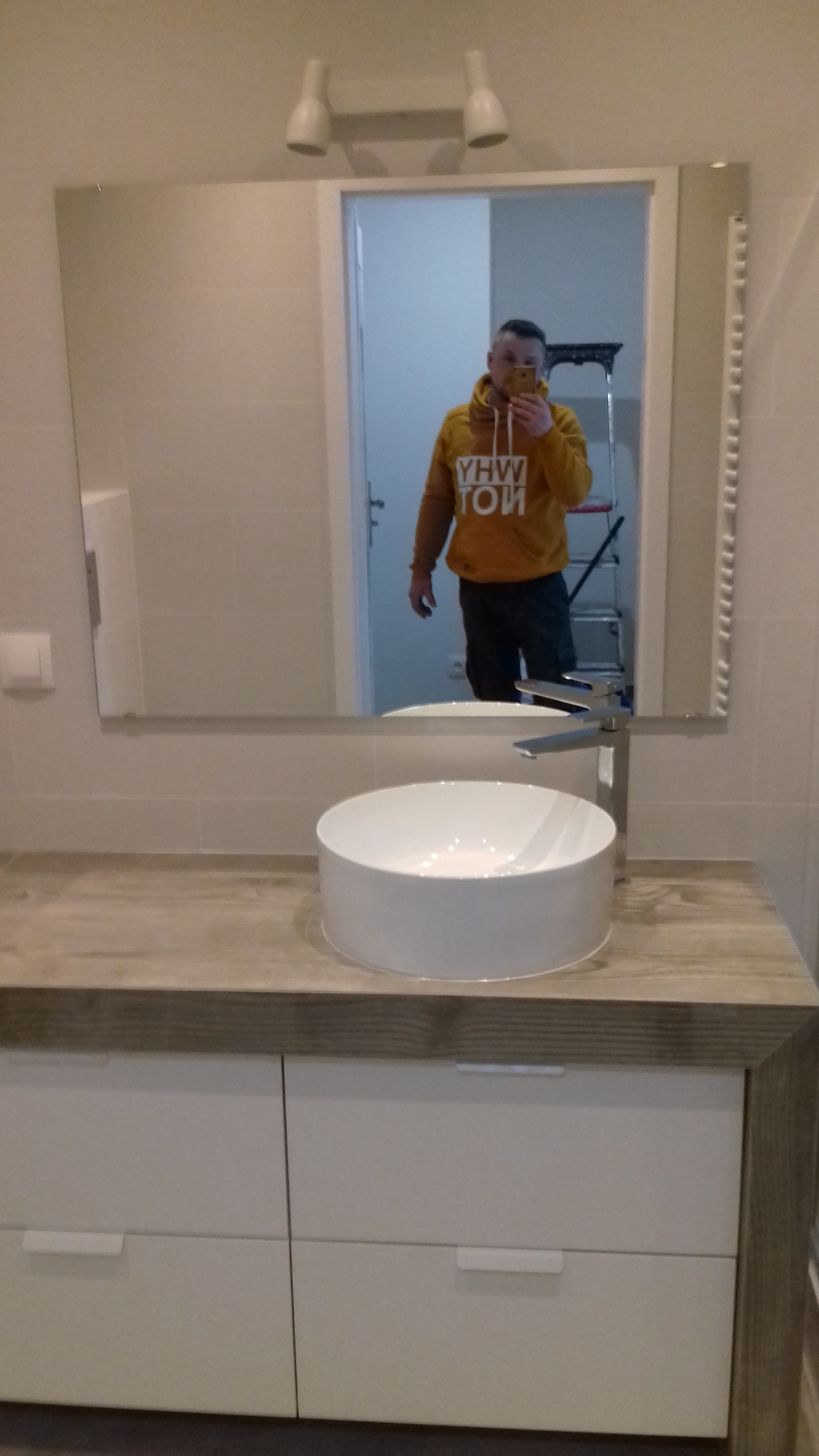 Nowoczesna łazienka z okrągłą umywalką nablatową, drewnopodobnym blatem i szafką z białymi frontami, odbicie osoby robiącej zdjęcie w lustrze, drabina w tle.