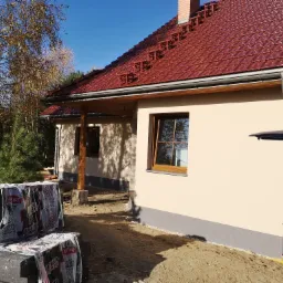 Nowo ocieplony dom jednorodzinny z brązową dachówką, widoczne materiały izolacyjne przygotowane do dalszych prac, jesienne drzewa w tle.