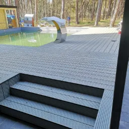 Kompozytowy taras z trzema schodkami prowadzącymi do basenu z ozdobną fontanną w kształcie łuku, w tle las sosnowy.