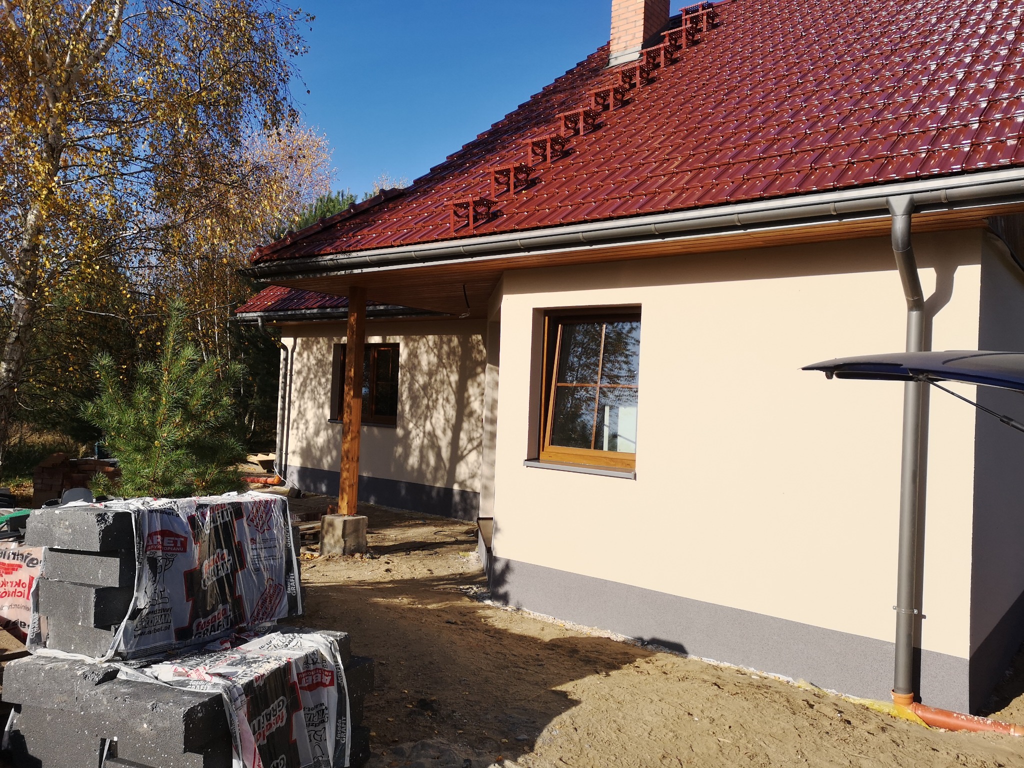 Nowo ocieplony dom jednorodzinny z brązową dachówką, widoczne materiały izolacyjne przygotowane do dalszych prac, jesienne drzewa w tle.