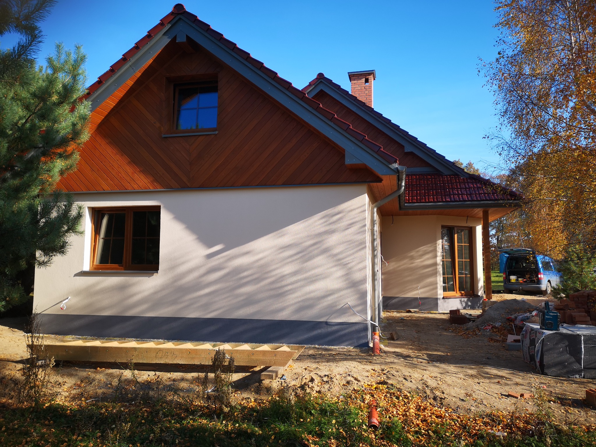 Nowo wybudowany dom jednorodzinny z drewnianą elewacją szczytową i jasną elewacją boczną, widoczne przygotowania do zagospodarowania terenu wokół budynku, częściowo ułożona kostka, jesienne liście...