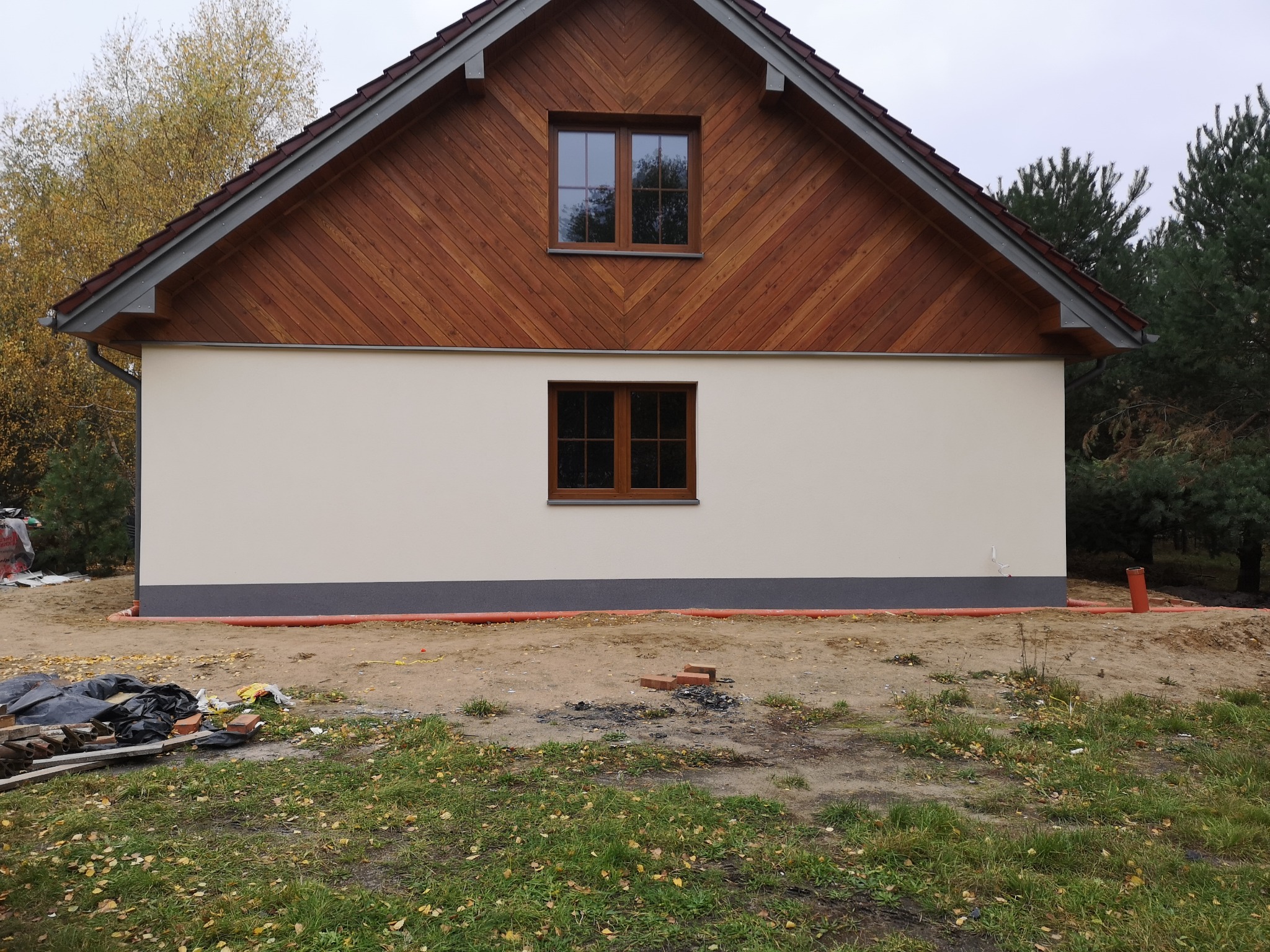 Wykończona elewacja domu jednorodzinnego z drewnianą górną częścią i dwoma oknami, widoczna pomarańczowa rura drenażowa przy fundamencie i nieporządek na trawniku.