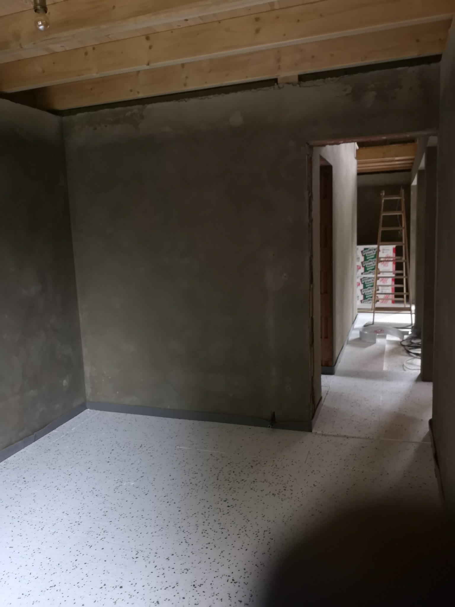 Surowe ściany po nałożeniu szpachli w nowym pokoju, widoczna drewniana konstrukcja sufitu, podłoga wyłożona jasną posadzką z ciemnymi plamkami, w głębi pomieszczenia oparta o ścianę drabina...