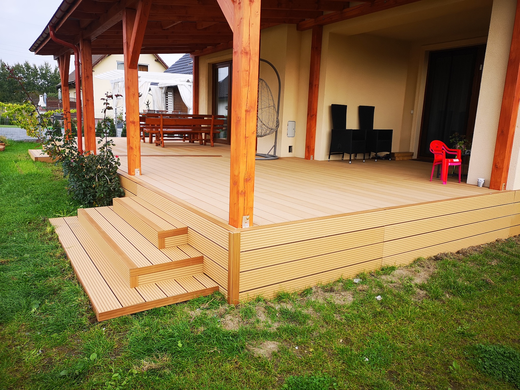 Zadaszony taras z desek kompozytowych z trzema schodkami na trawnik, drewniana konstrukcja dachu, w tle dom z pergolą i huśtawką.
