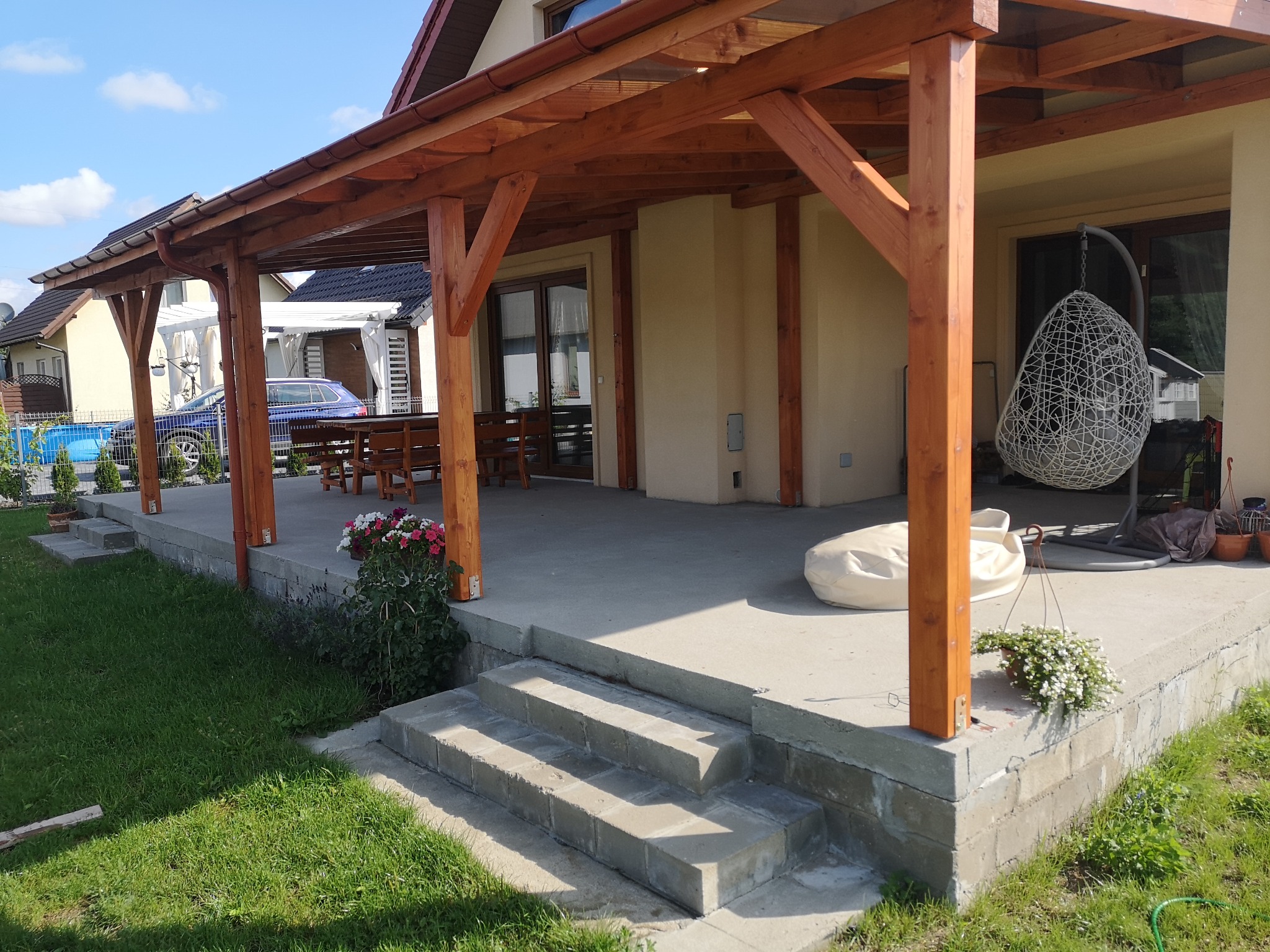 Zadaszony taras z drewnianą konstrukcją, betonową wylewką i schodami, widok na ogród z meblami ogrodowymi i wiszącym fotelem.