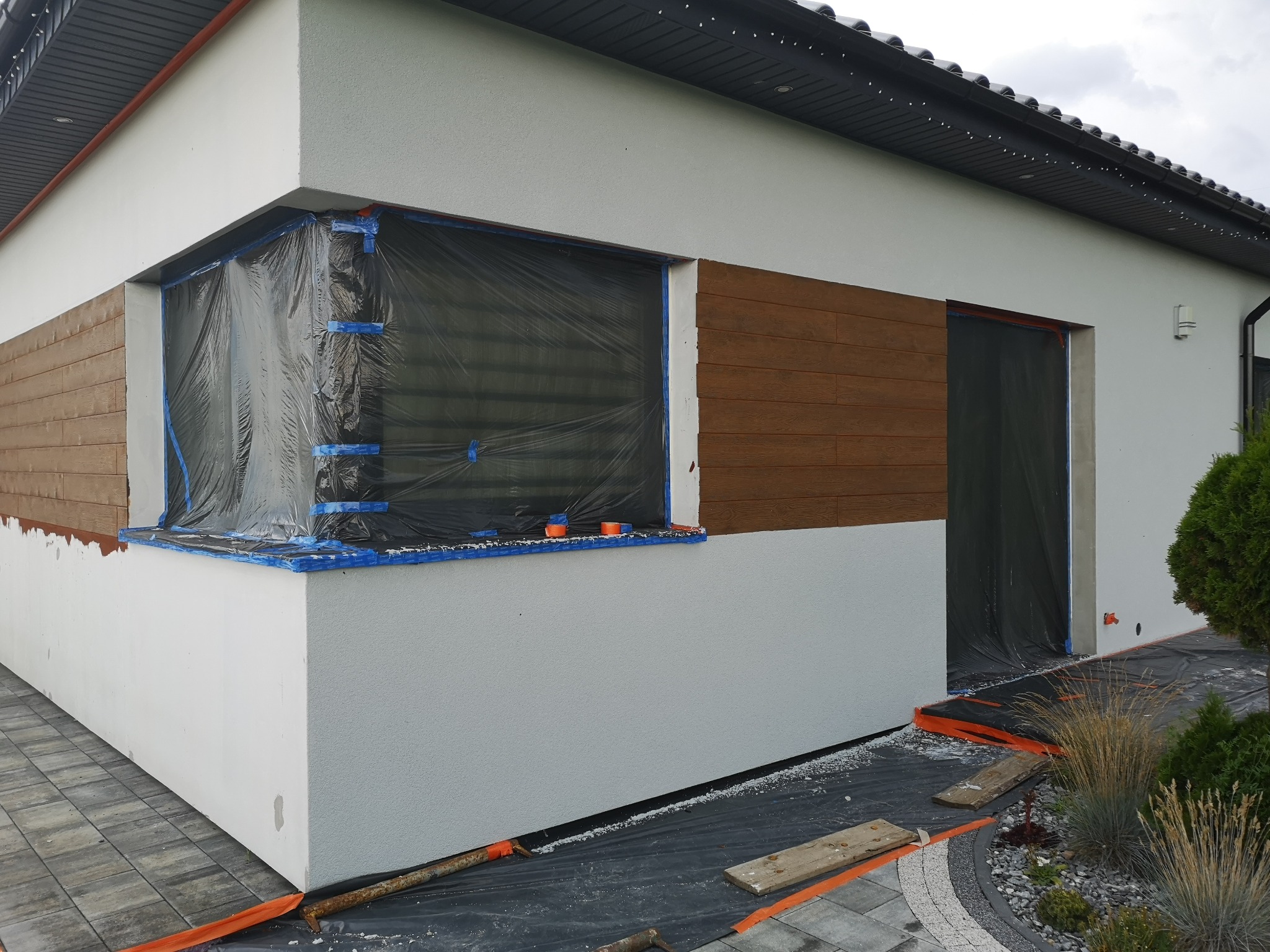 Przygotowanie elewacji budynku do montażu drewnianej okładziny, okna zabezpieczone folią, widoczne fragmenty ocieplonej ściany i ułożonej kostki brukowej.