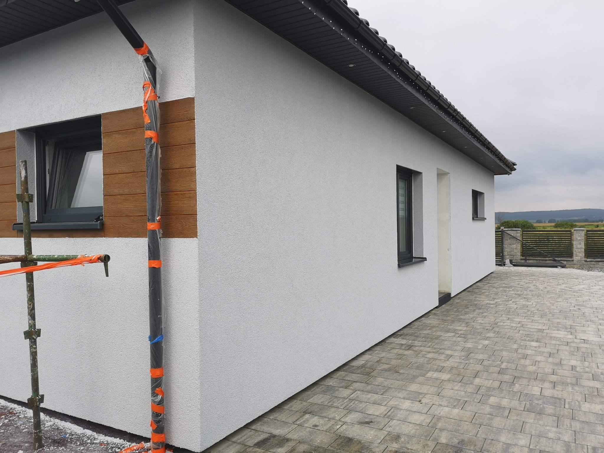 Świeżo otynkowana elewacja domu jednorodzinnego z drewnopodobną okładziną, zabezpieczona rynna w folii i rusztowanie obok okna.