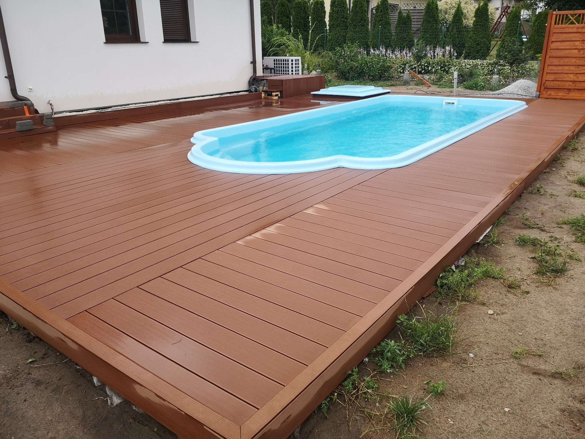 Niebieski basen z tworzywa sztucznego otoczony brązowym tarasem z desek kompozytowych, widok z poziomu gruntu.