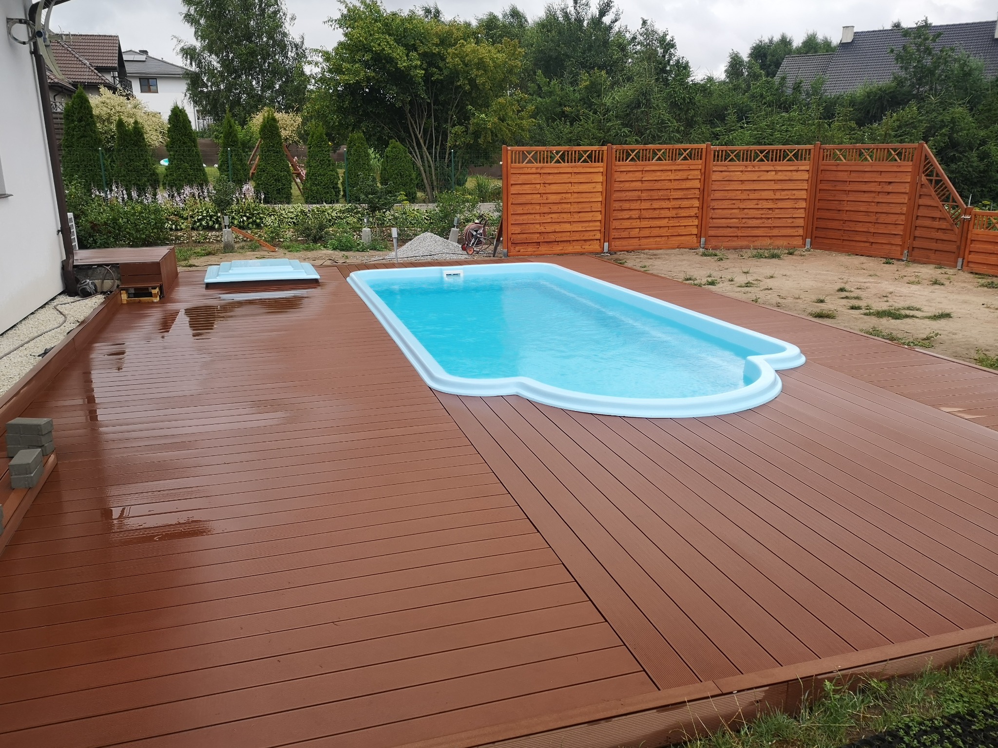 Nowoczesny basen ogrodowy z jasnoniebieską wodą, otoczony brązowym tarasem kompozytowym i drewnianym ogrodzeniem, widok z góry w pochmurny dzień.