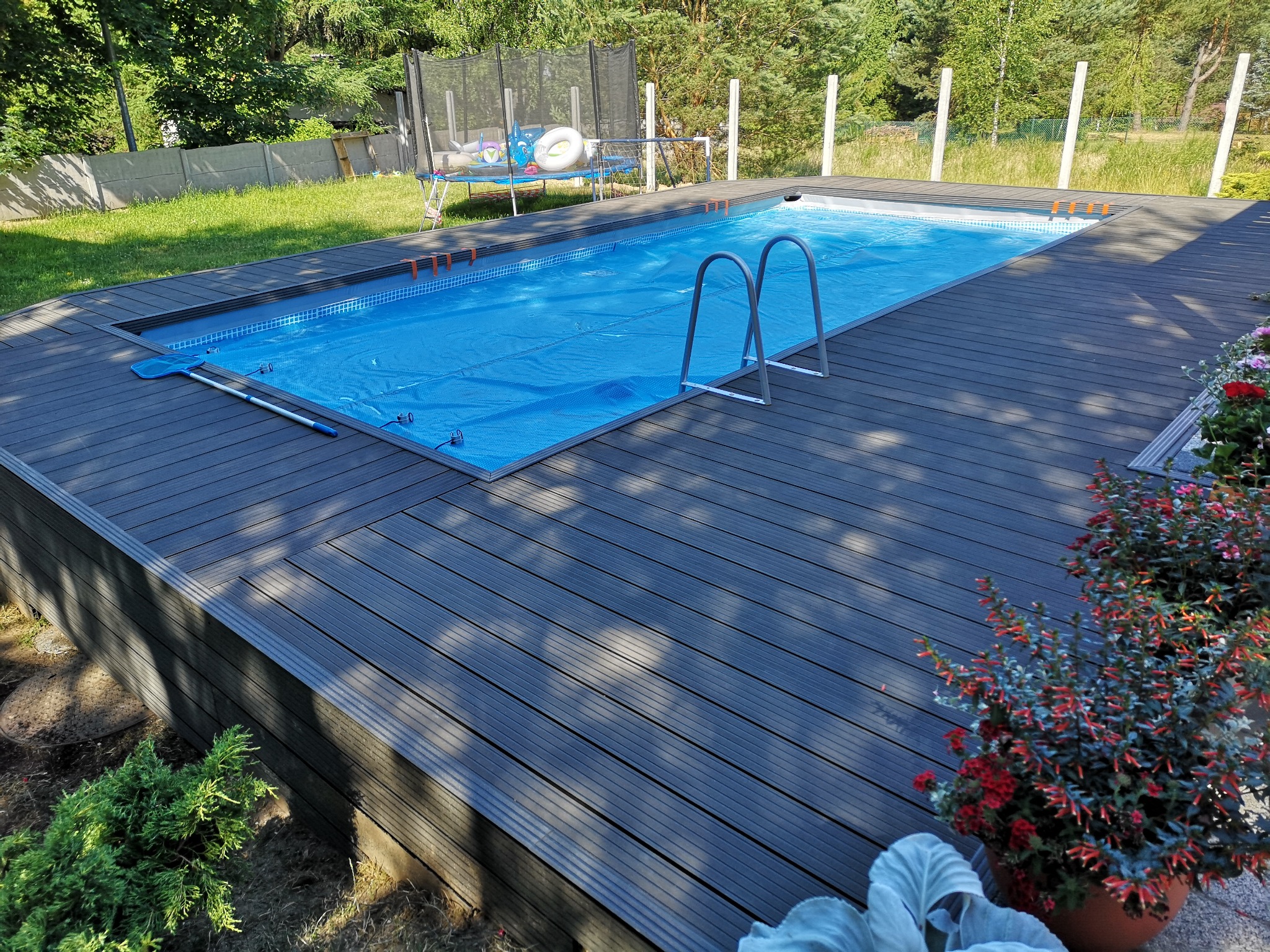 Zadaszony basen ogrodowy z drabinką, otoczony ciemnoszarą, kompozytową deską tarasową, na tle zieleni drzew i trawnika.