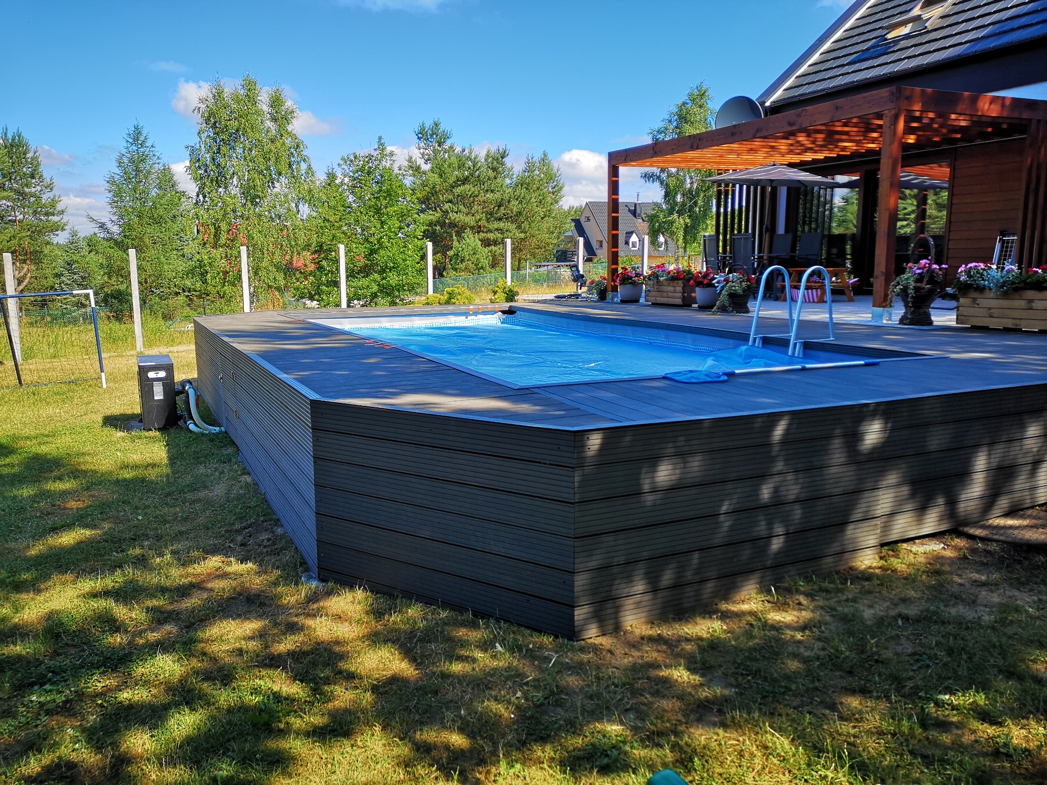 Zewnętrzny basen z szarą obudową z desek kompozytowych, przykryty niebieską folią, z metalową drabinką, otoczony zielonym trawnikiem, w tle drzewa i pergola przy budynku.