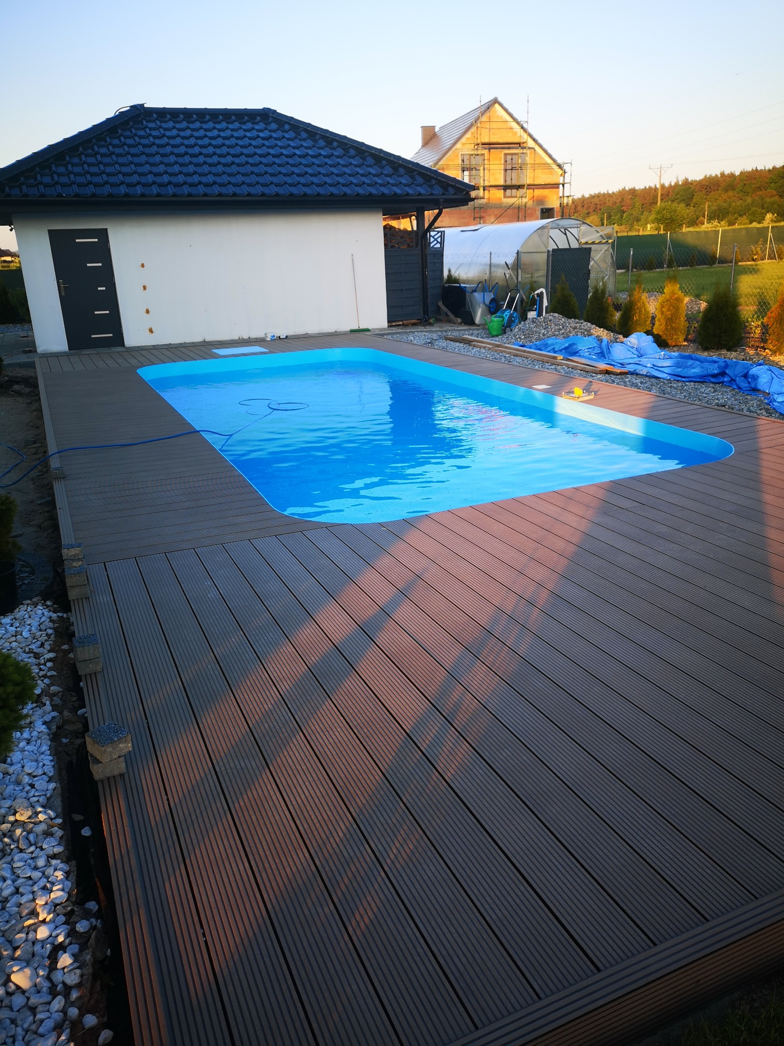 Niebieski basen otoczony tarasem z desek kompozytowych, widok na budynek gospodarczy i dom w trakcie budowy, w tle zieleń.