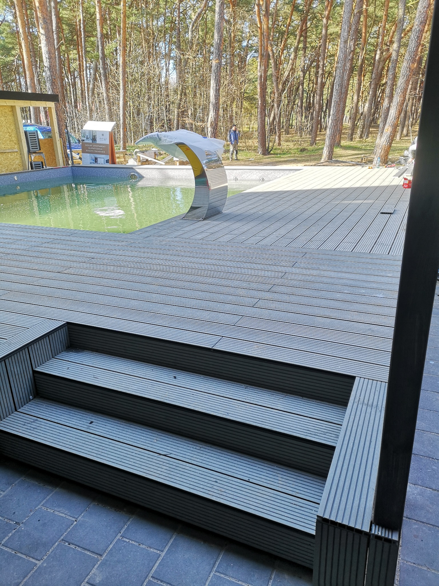 Kompozytowy taras z trzema schodkami prowadzącymi do basenu z ozdobną fontanną w kształcie łuku, w tle las sosnowy.