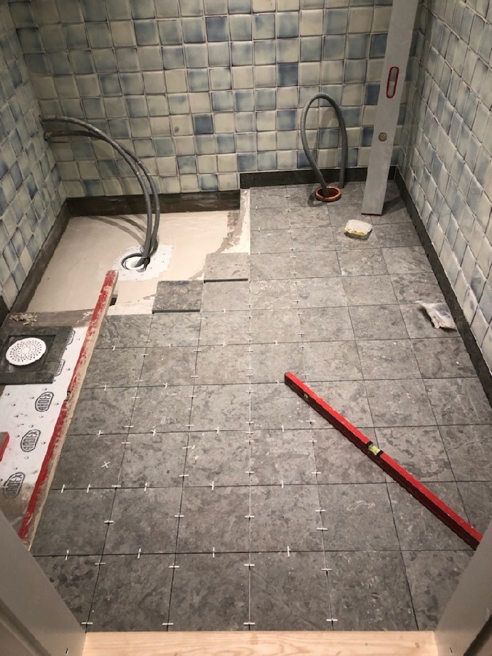 Łazienka w trakcie układania szarych płytek podłogowych, widoczne krzyżyki dystansowe, poziomica, fragment podłoża z wylewką i odpływem liniowym, niebiesko-białe płytki na ścianach.